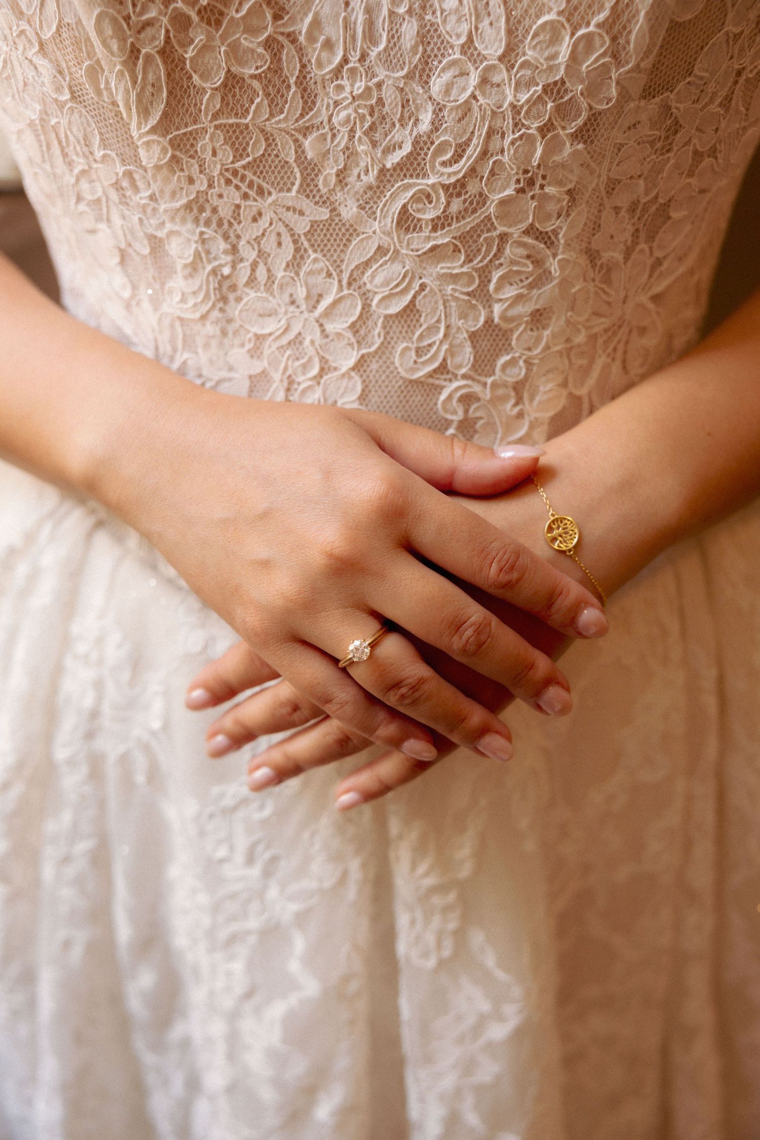 Bride in lace wedding dress with hands clasped behind back, wearing diamond engagement ring and gold signet ring.
