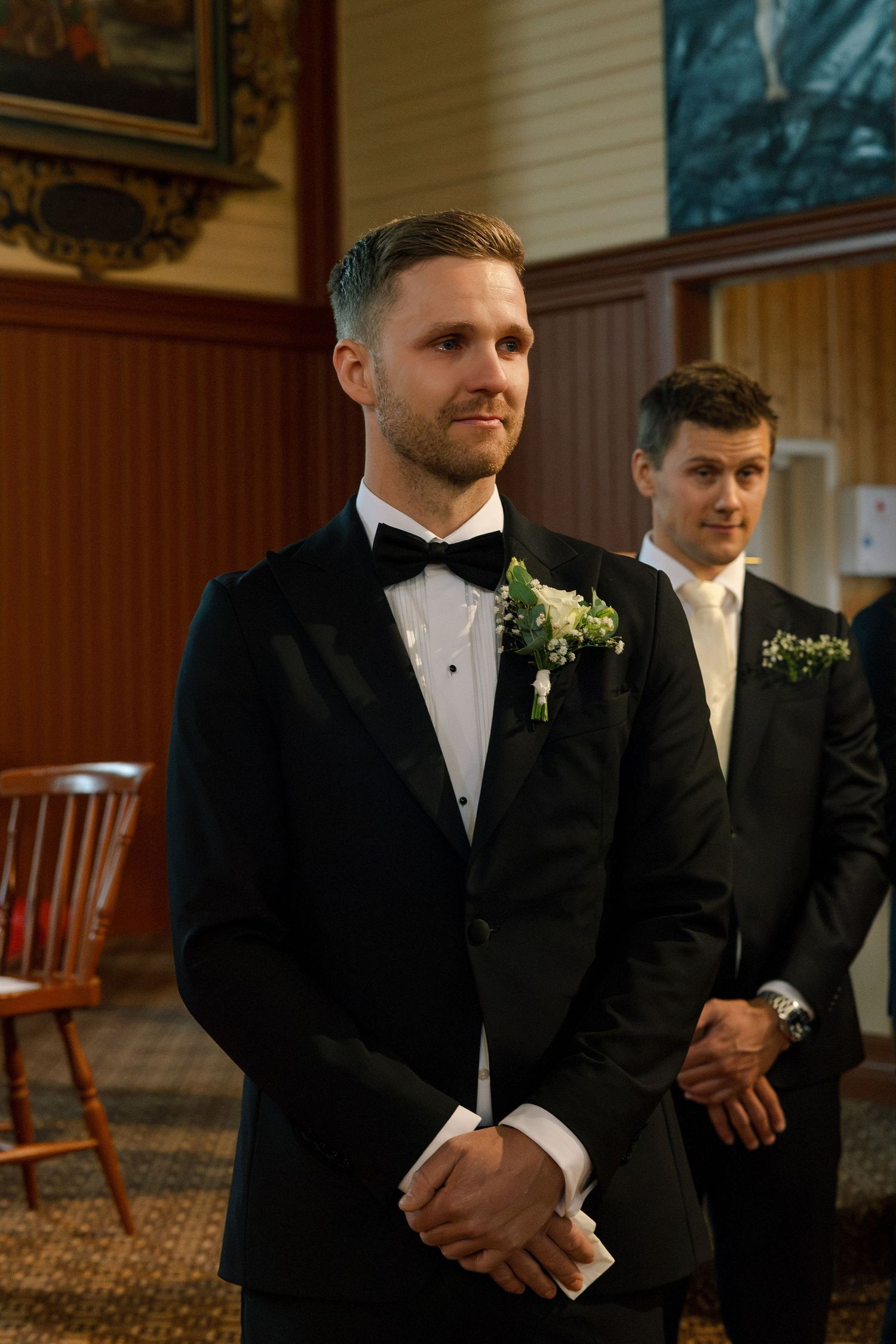 Groom in black tuxedo with boutonniere stands at altar, groomsman visible behind him during wedding ceremony.