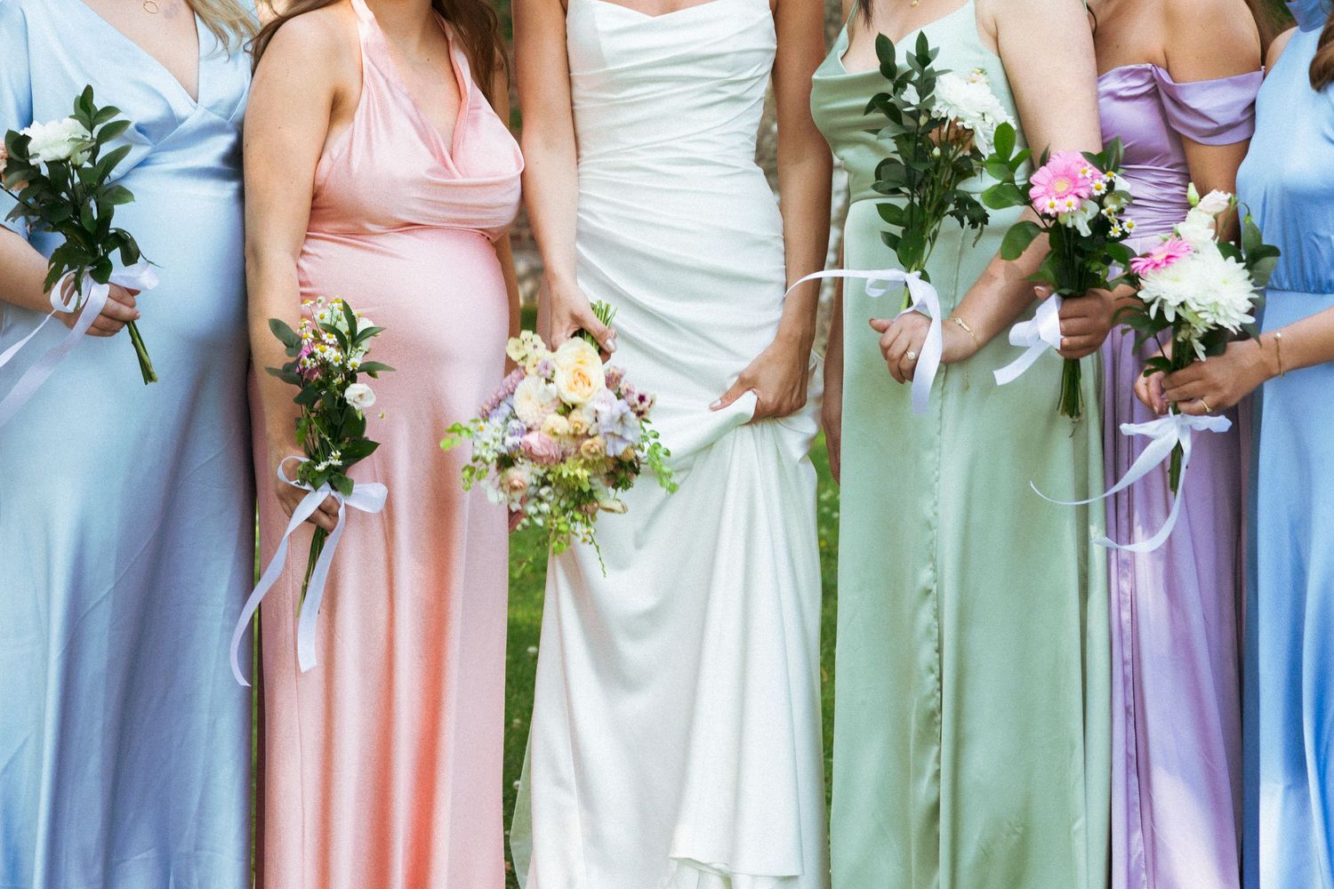Bride in white gown poses with six bridesmaids in pastel multicolored dresses holding floral bouquets outdoors.