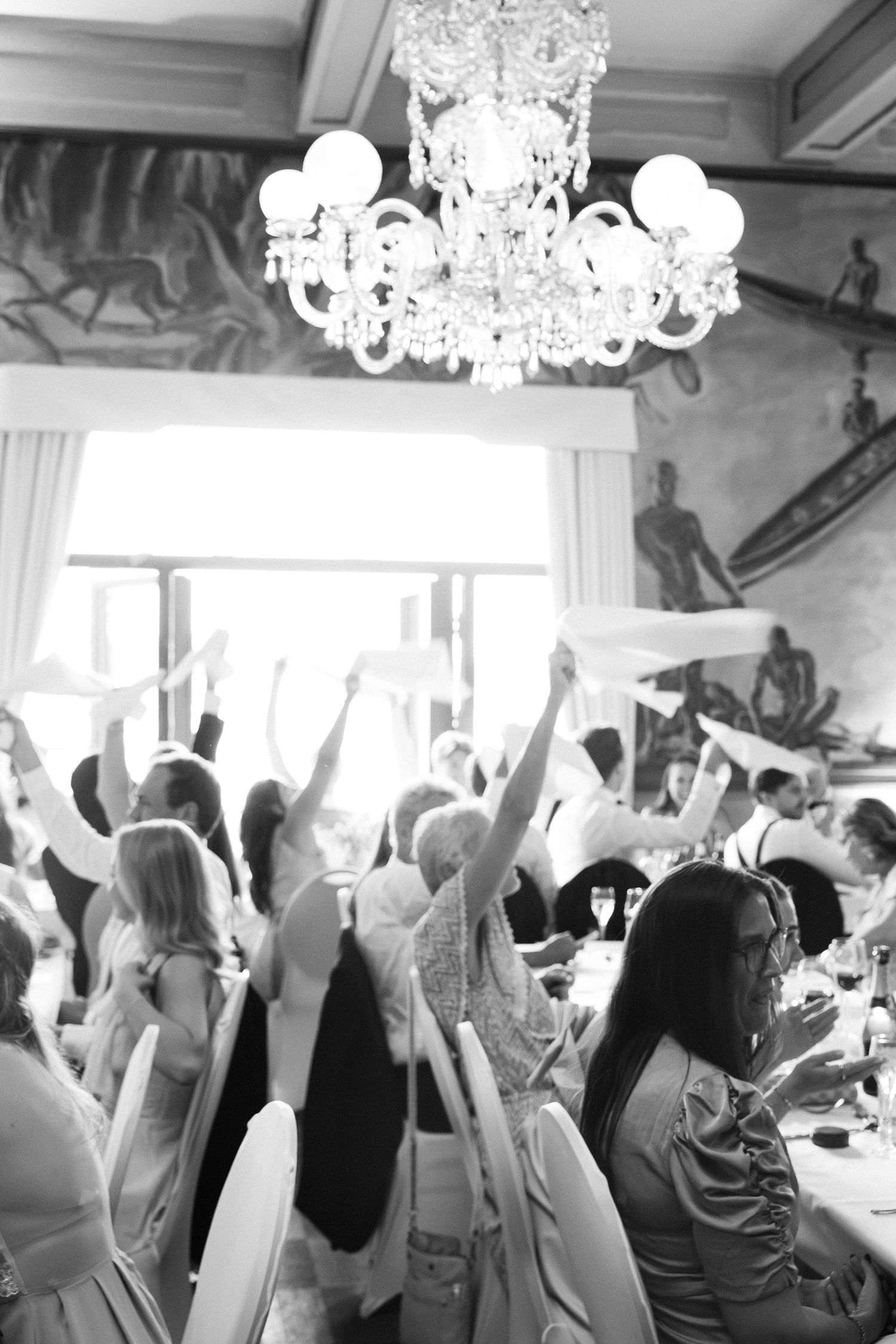 Wedding guests wave napkins under crystal chandelier in elegant ballroom.