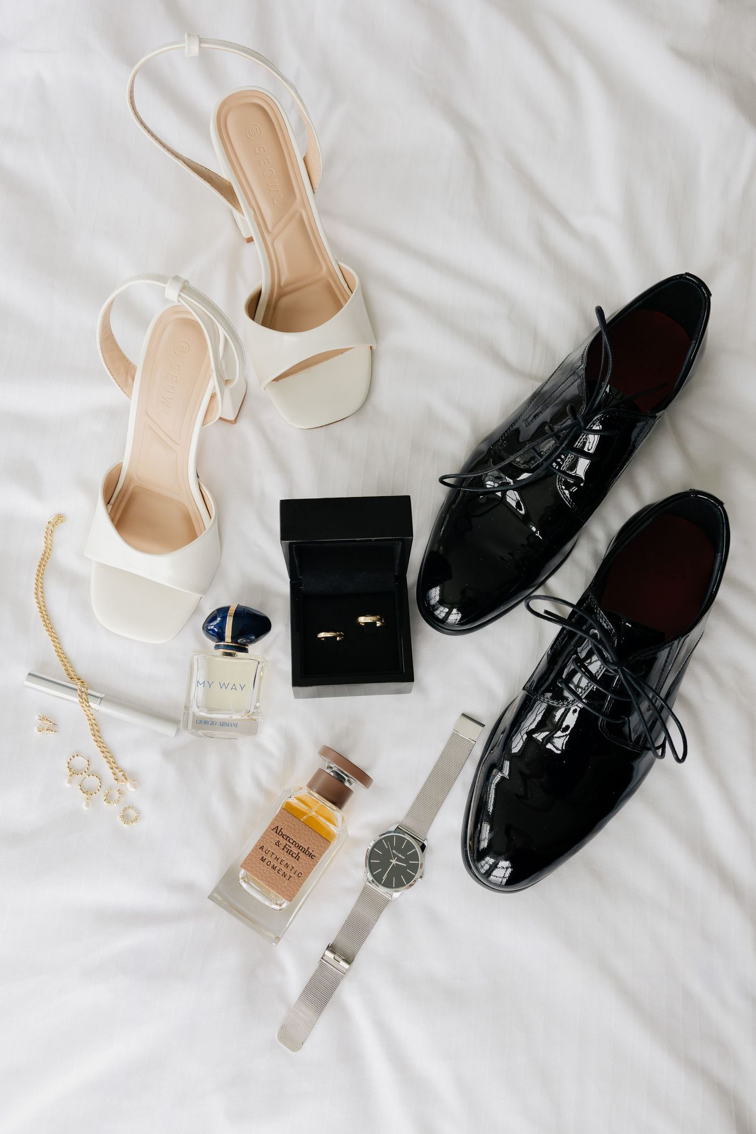 Wedding accessories flat lay featuring white heels, black patent dress shoes, rings, watches, perfume, and pearl jewelry on white fabric.