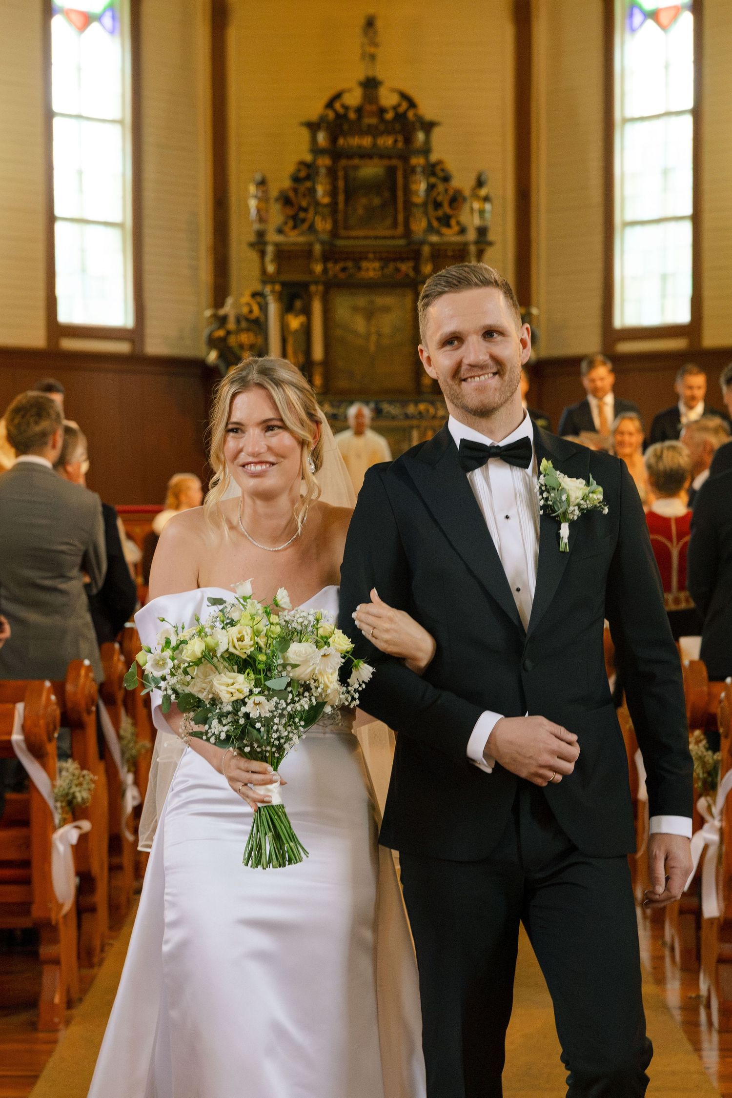 Bride holding white bouquet and groom in tuxedo walking down church aisle after ceremony.