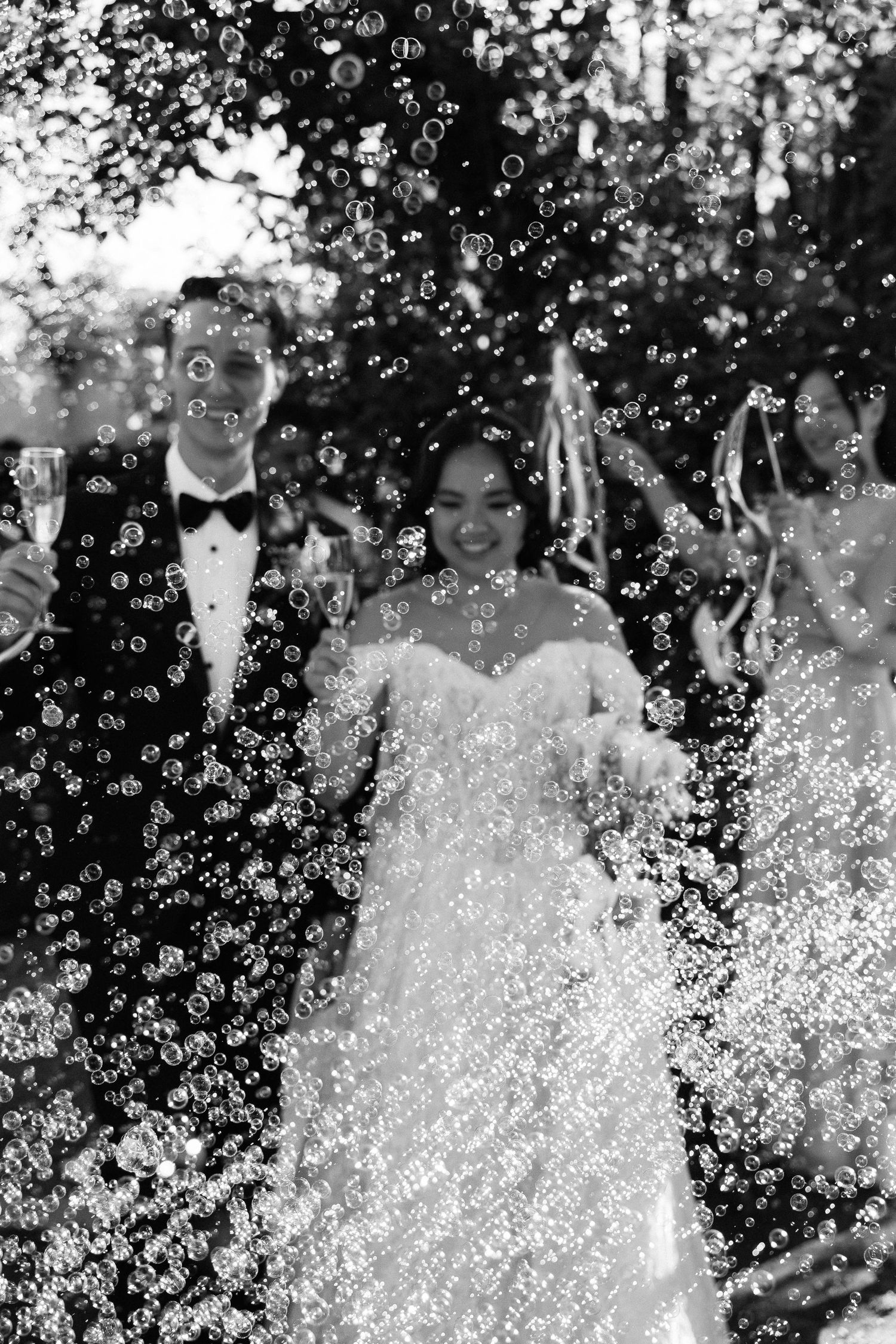 Black and white wedding photo of smiling bride and groom surrounded by floating bubbles outdoors.