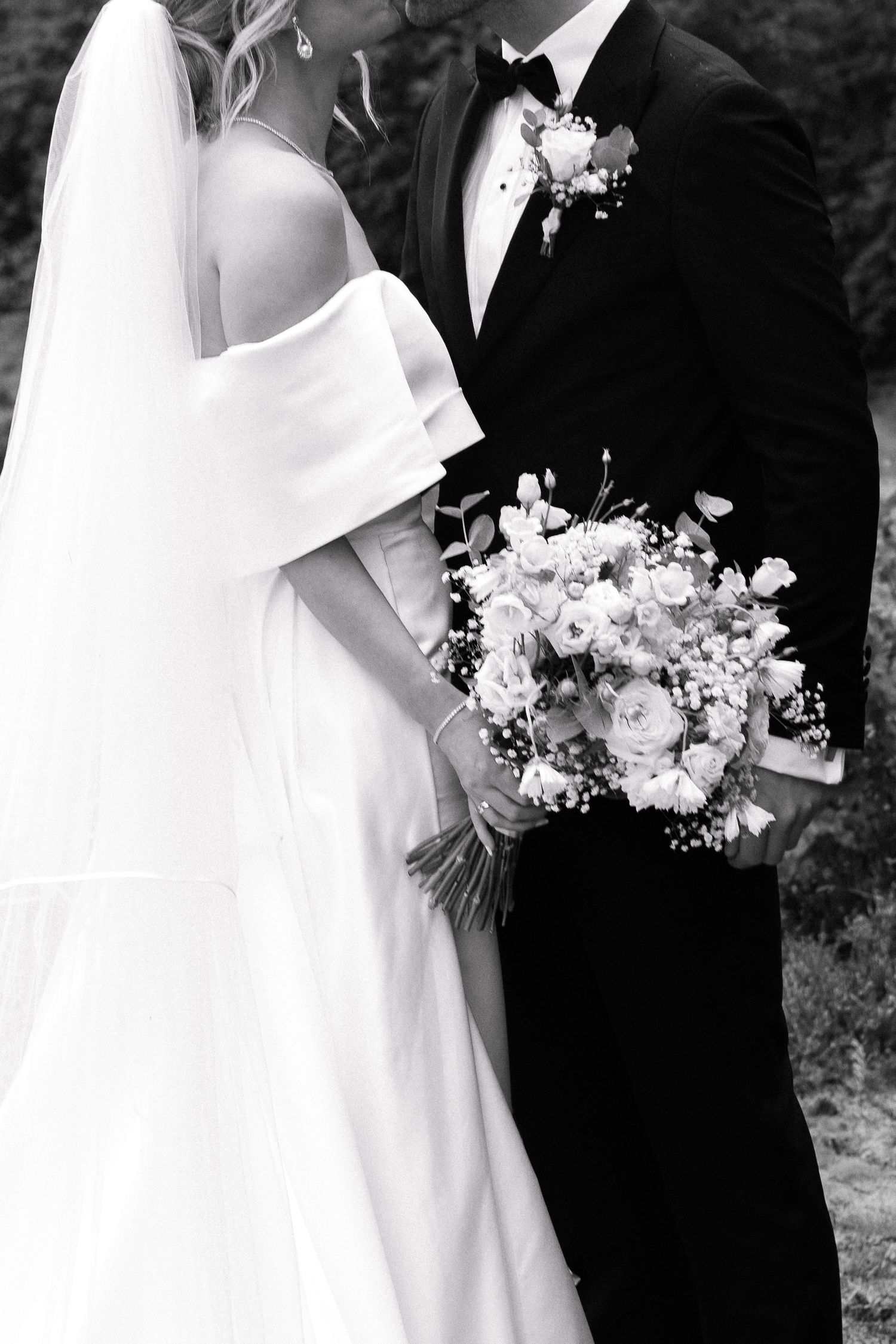 Black and white portrait of bride in off-shoulder gown and groom in tuxedo holding floral bouquet.