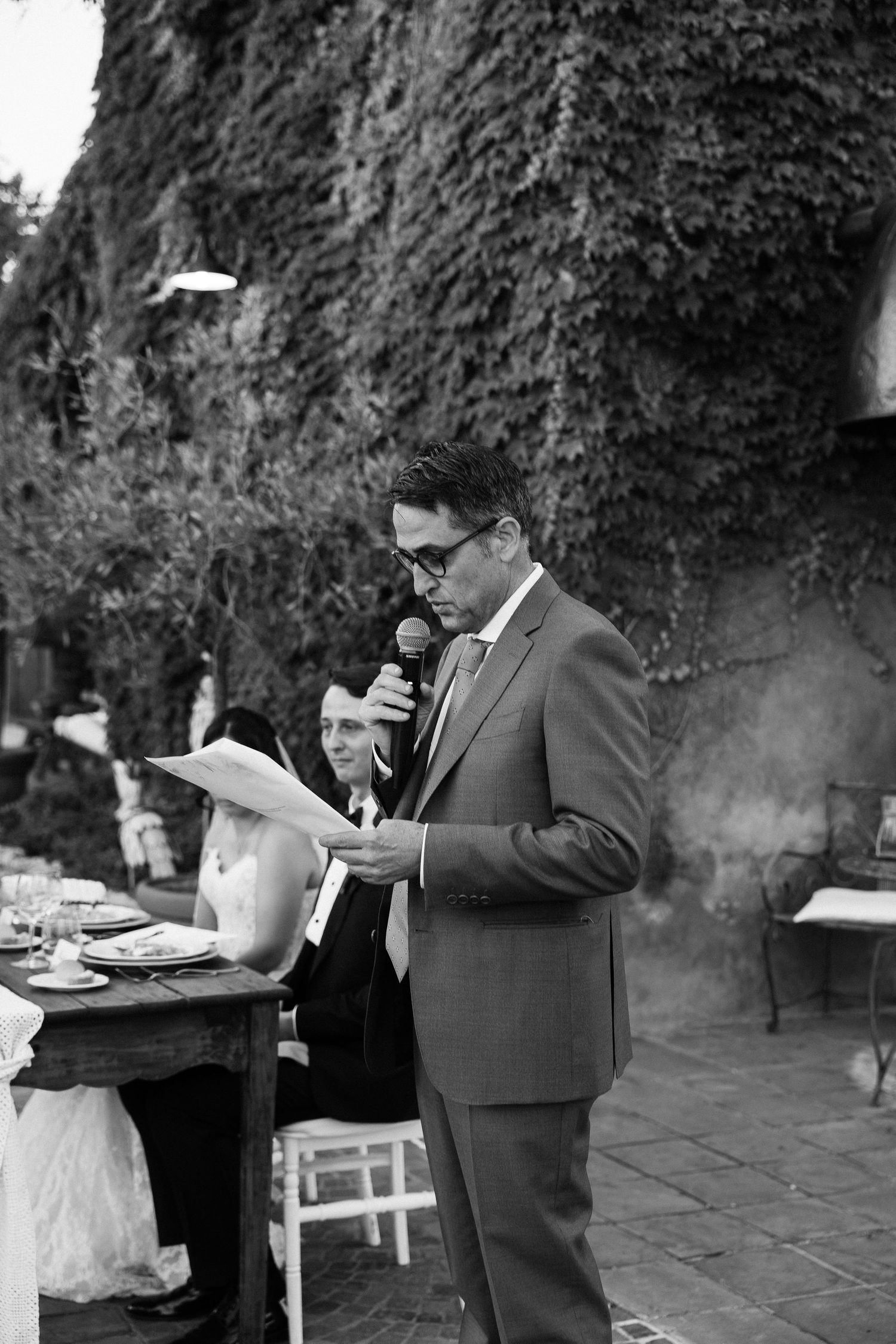 Black and white photo of groomsman delivering speech at outdoor wedding reception holding microphone.