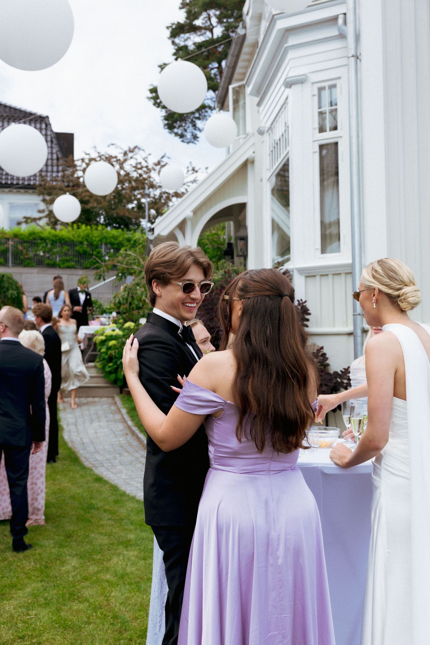 Groom in black tuxedo with sunglasses chatting with guests in purple dress at outdoor wedding.