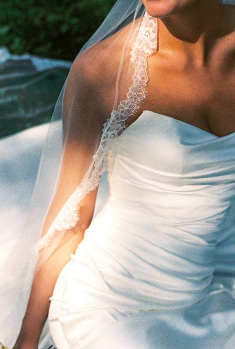 Close-up of bride in strapless ruched white wedding gown with delicate lace-trimmed veil in warm sunlight.