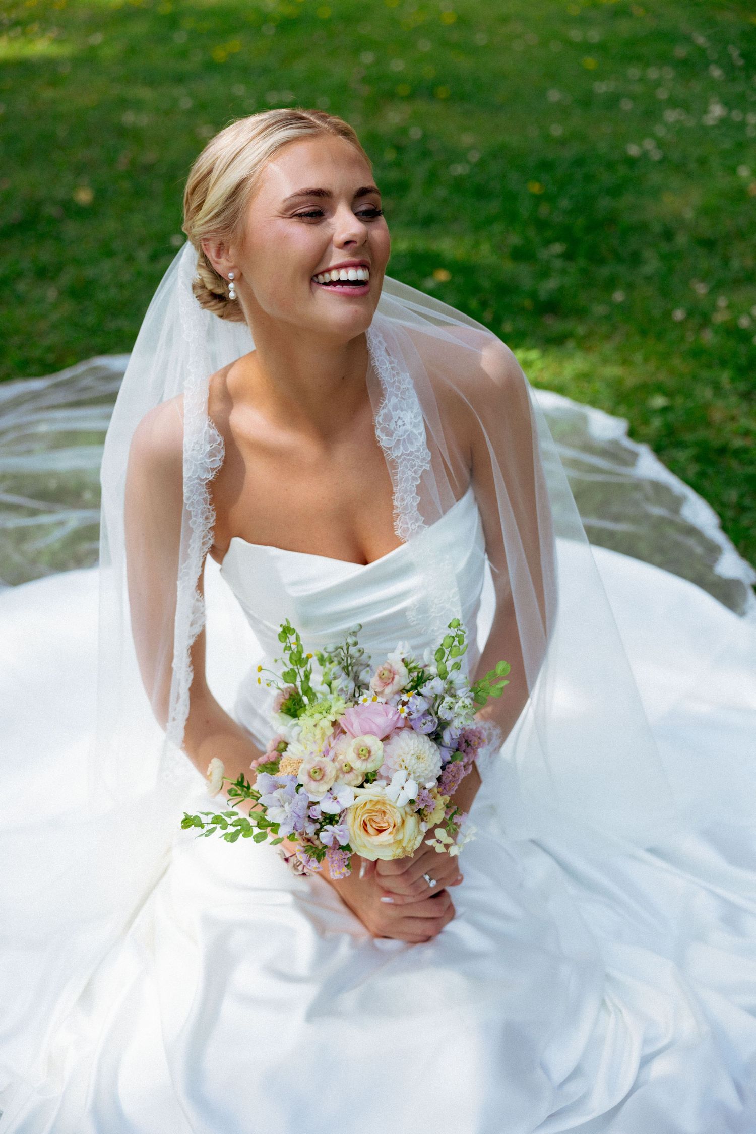 Happy blonde bride in white gown sitting on grass holding pastel flower bouquet.