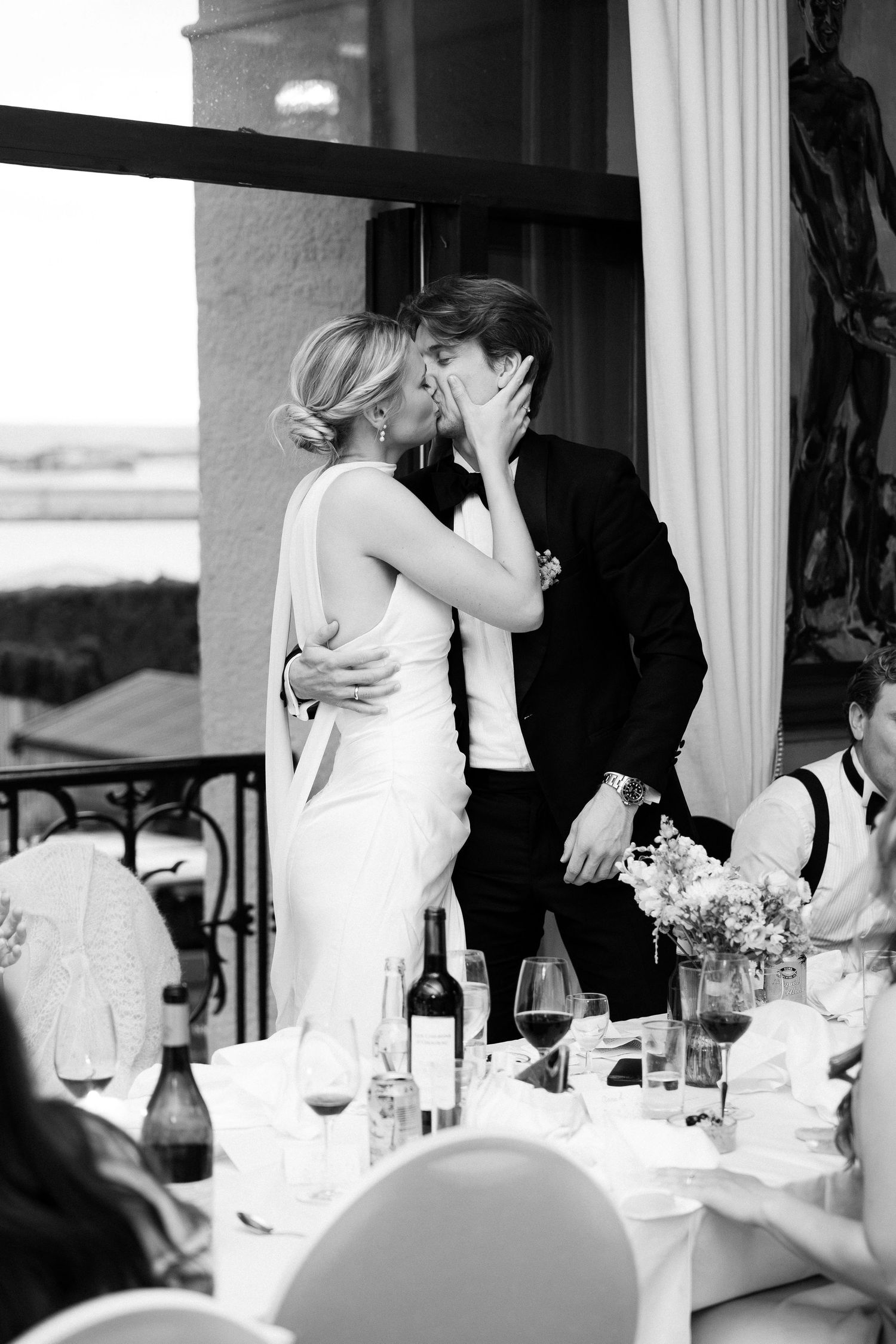 Black and white photo of bride and groom sharing a kiss at wedding reception dinner table.