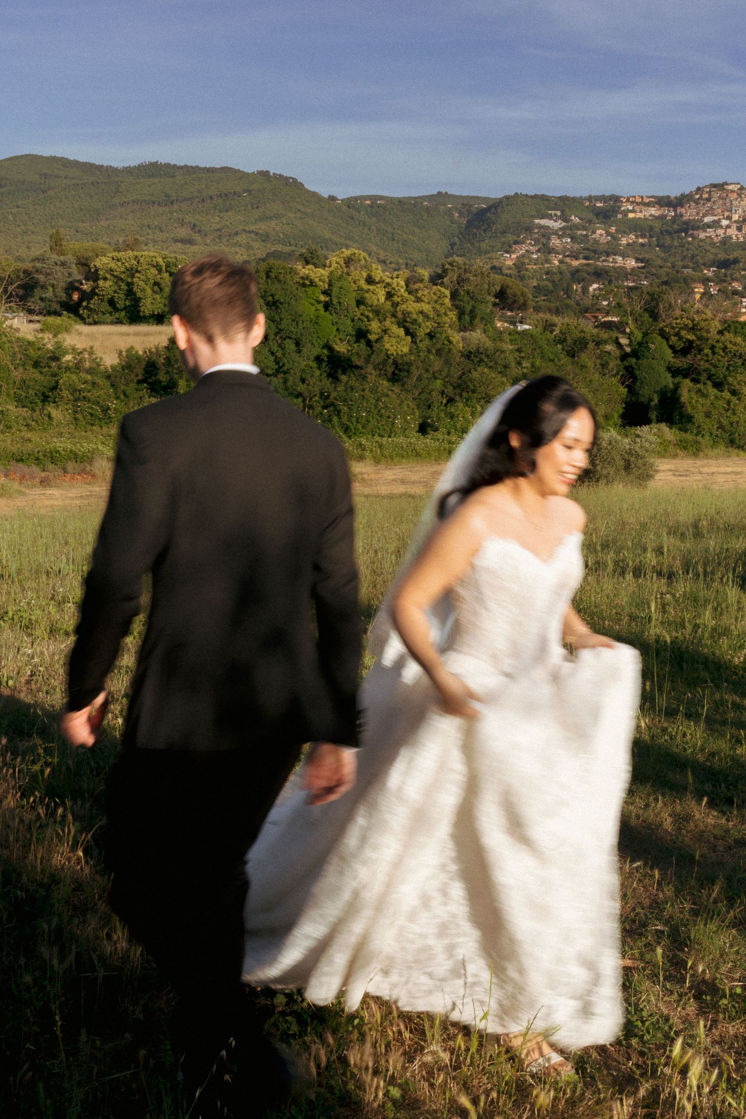 Bride and groom walking through green meadow with mountain backdrop on sunny wedding day.