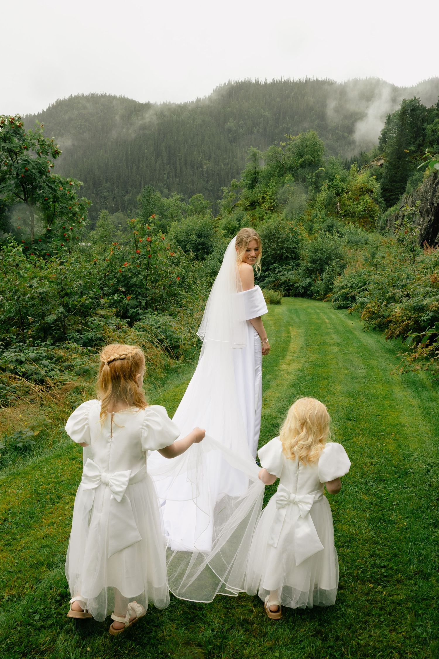 Bride in white gown and veil walks with two flower girls on lush green path with misty mountains.