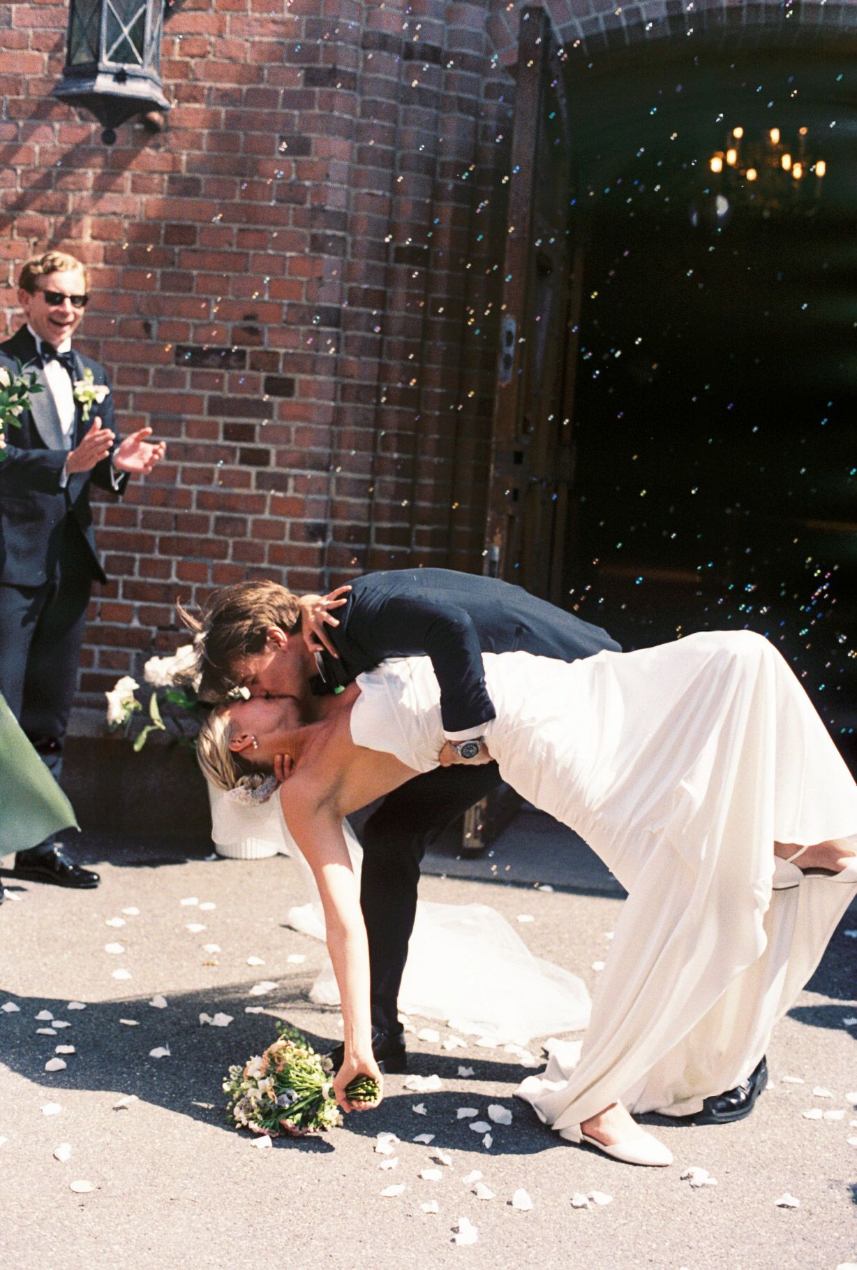 Bride and groom share a romantic dip kiss outside brick church, with confetti and guests celebrating their wedding day.