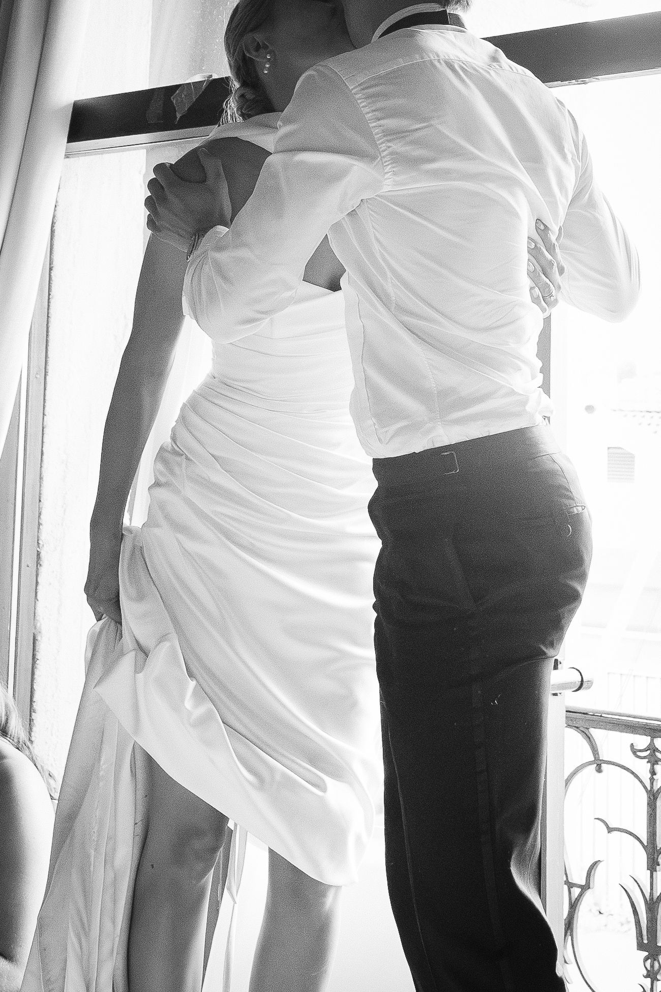 Black and white photo of bride and groom embracing by a window in wedding attire.