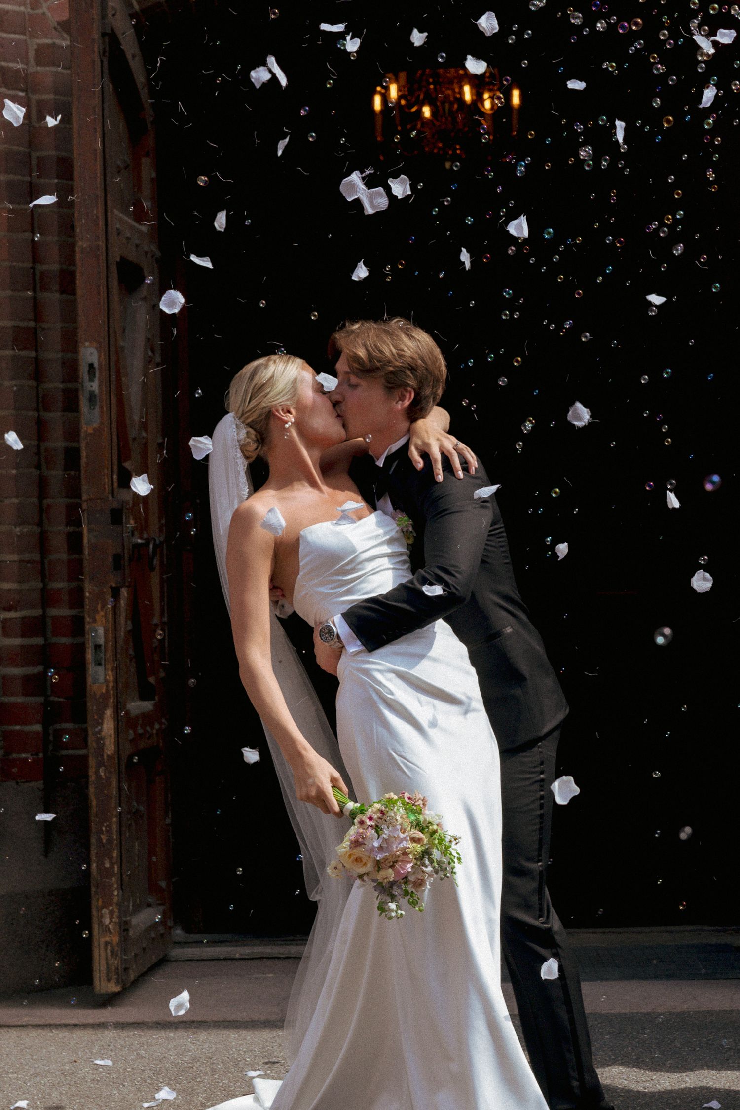 Bride and groom kissing outside church entrance with white confetti falling around them.