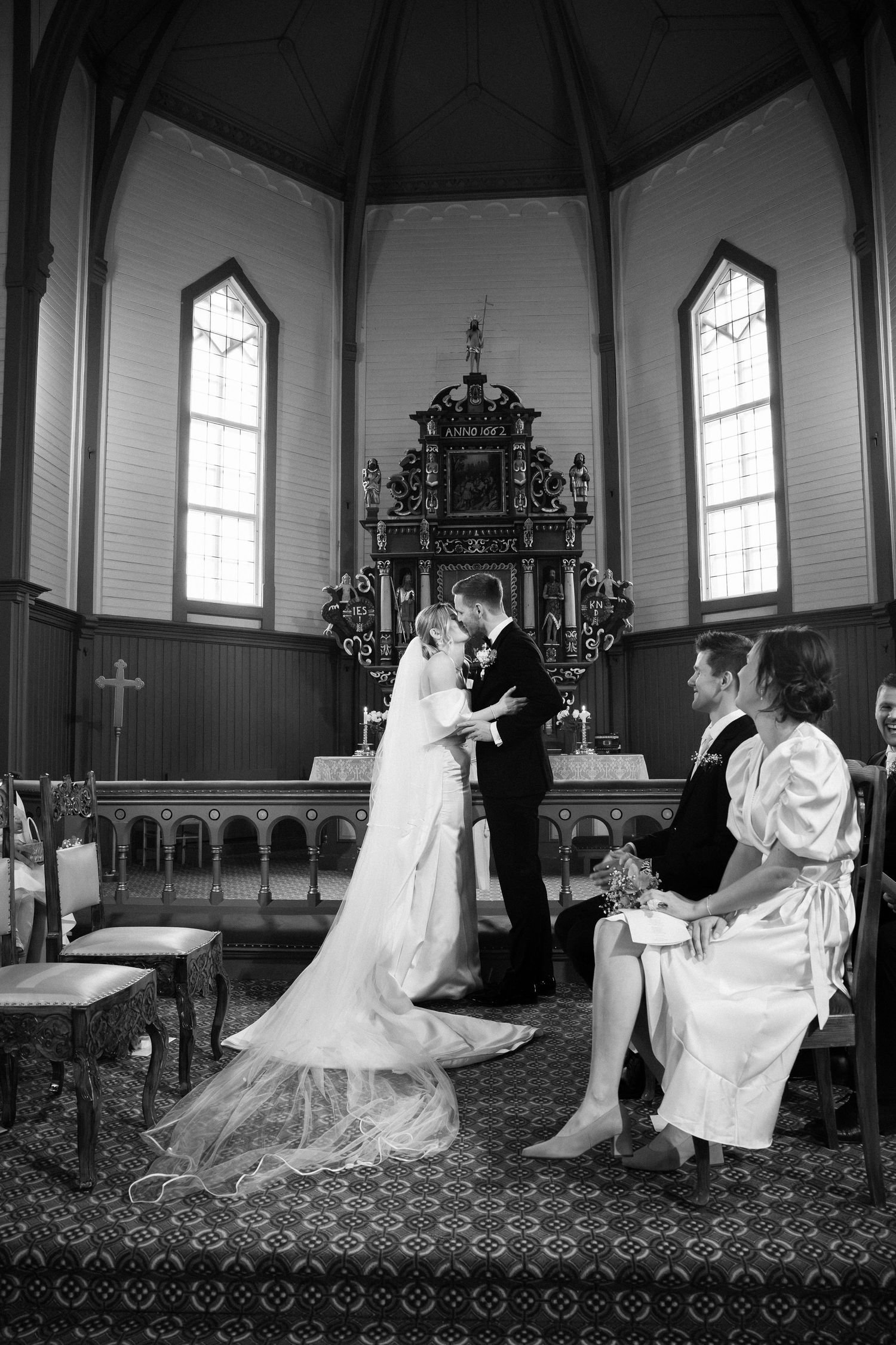 Black and white photo of bride and groom exchanging vows at a church altar with guests seated nearby.