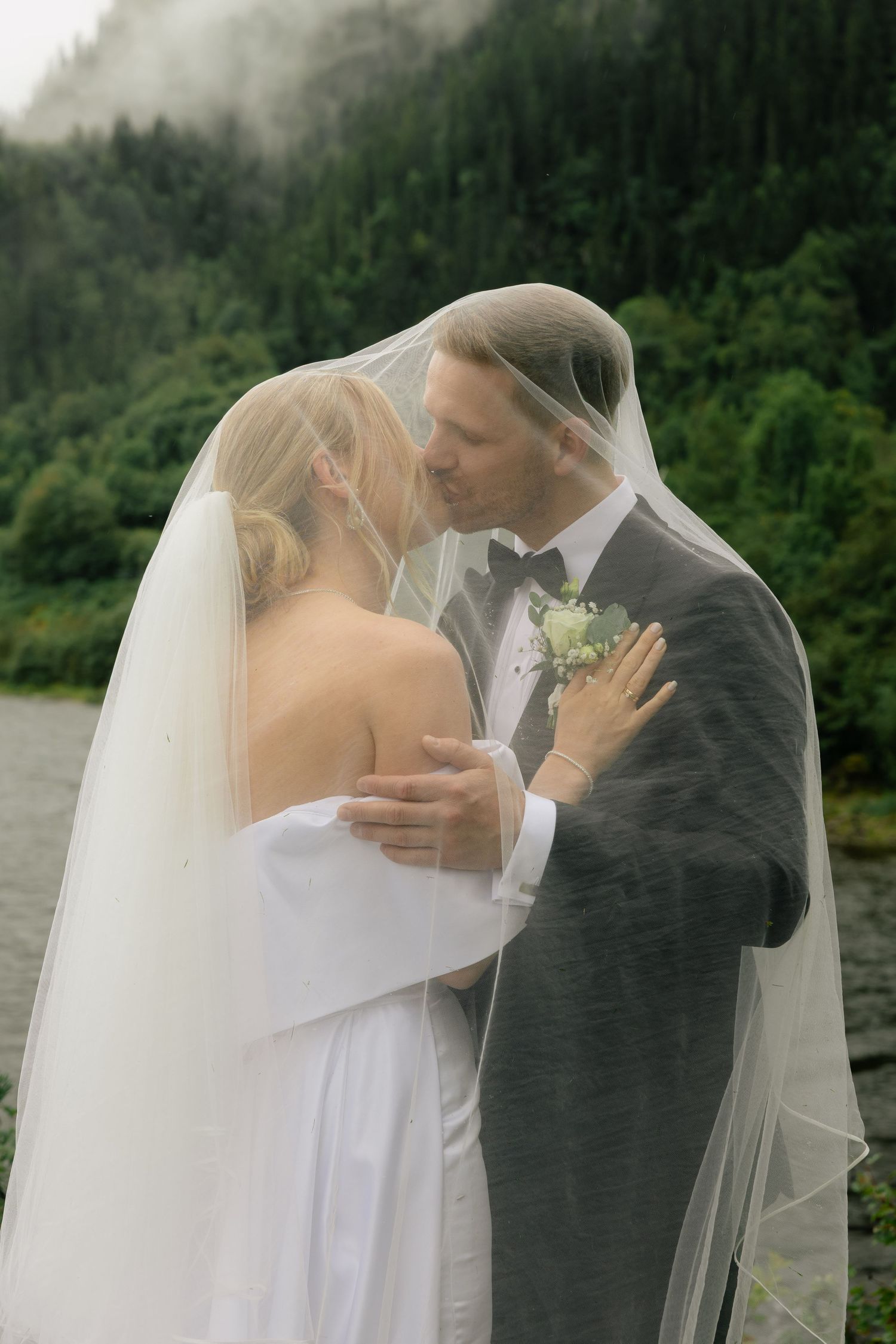 Bride and groom sharing romantic kiss under bridal veil with misty green forest and lake behind them.