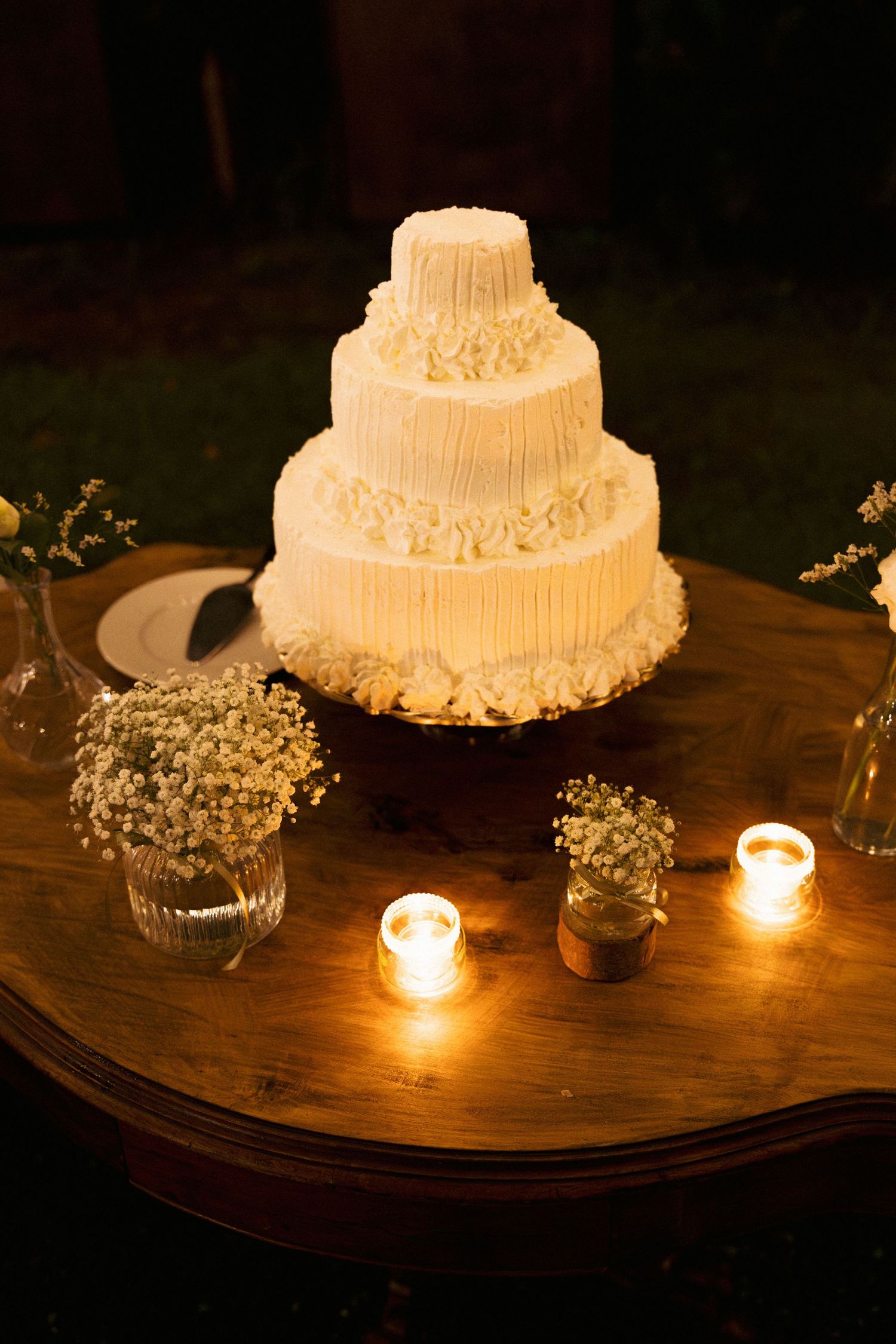 Elegant four-tier white wedding cake with floral details surrounded by candles and baby's breath on wooden table.