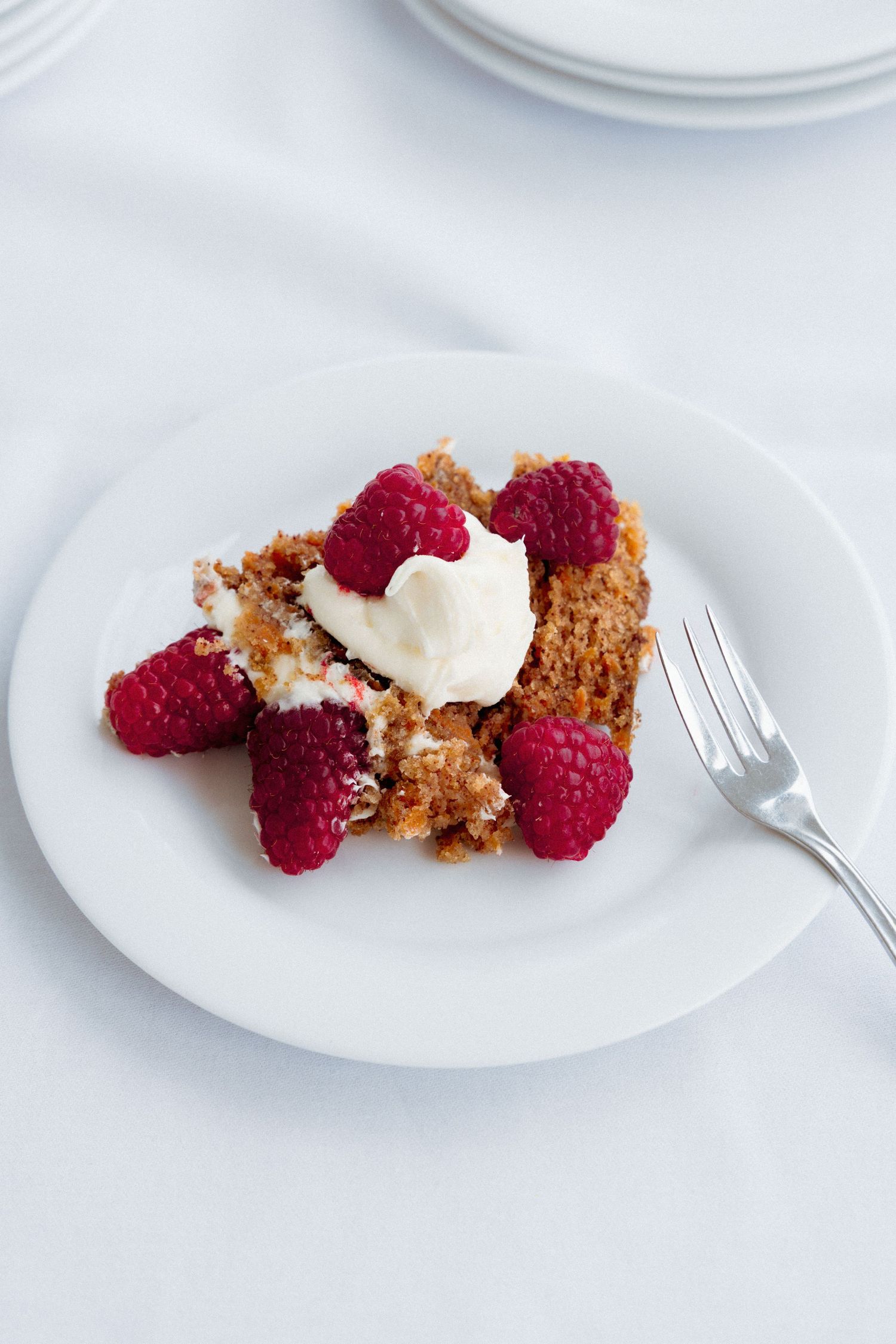 A white plate with a slice of cake topped with fresh raspberries and whipped cream, served with a fork on a white tablecloth.