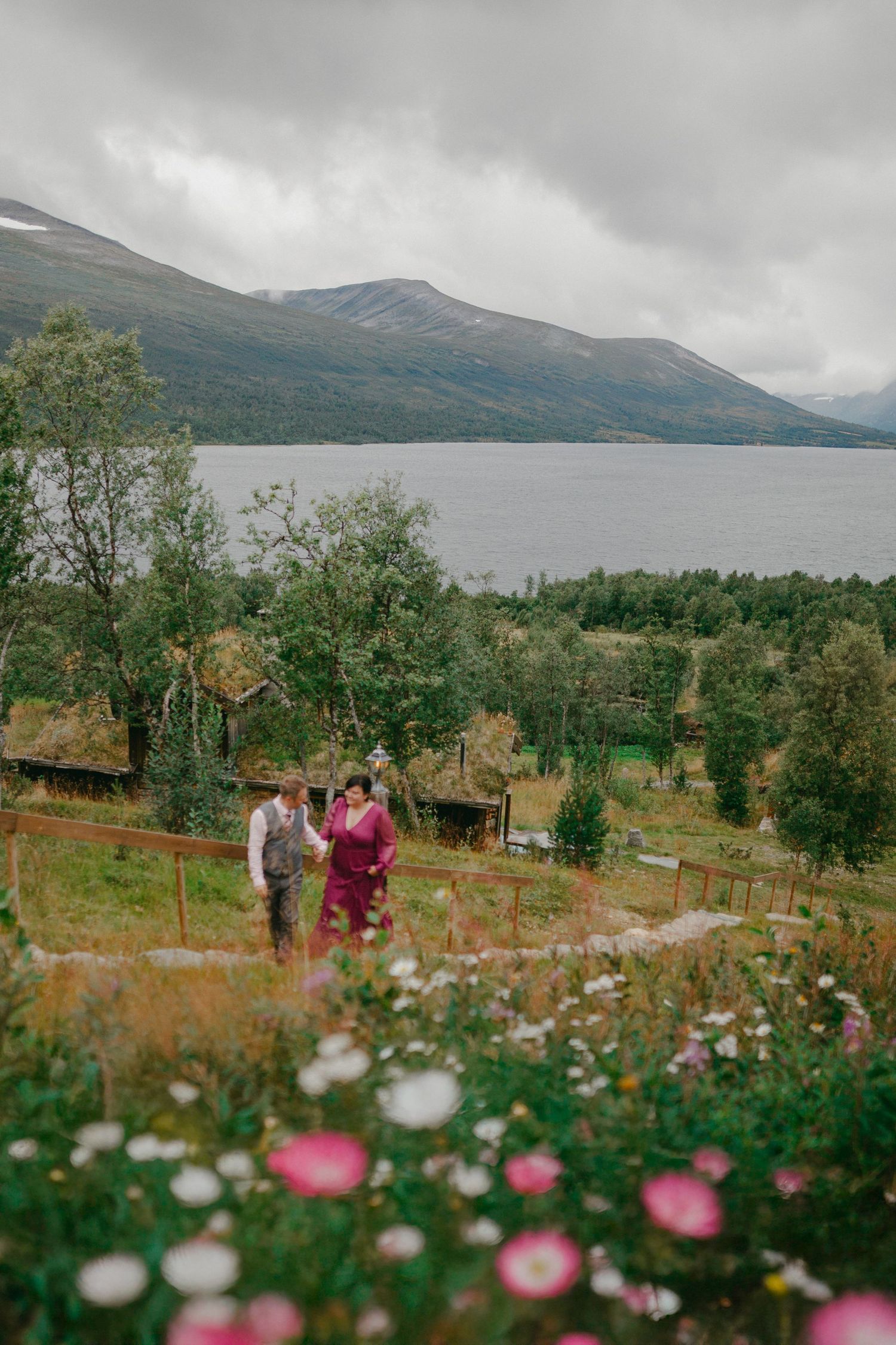 Couple walks through wildflower meadow with fjord and mountain backdrop on cloudy day.