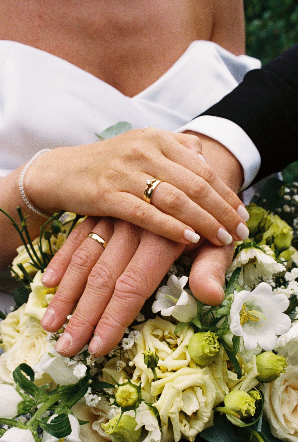 Newlyweds displaying gold wedding rings on hands resting over white bridal bouquet.