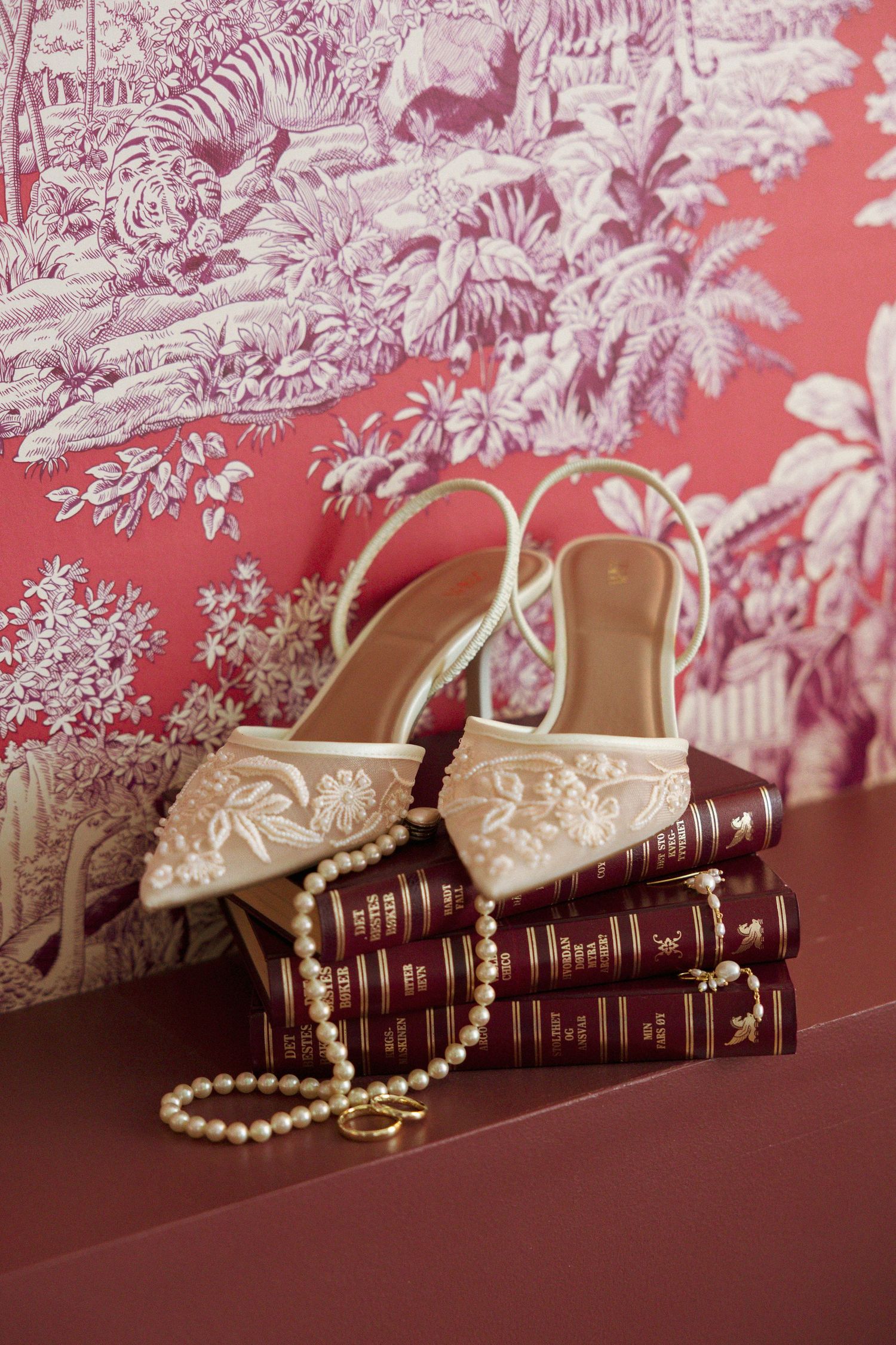 Elegant white embroidered heels resting on vintage books with pearl necklace, against pink toile de Jouy wallpaper.