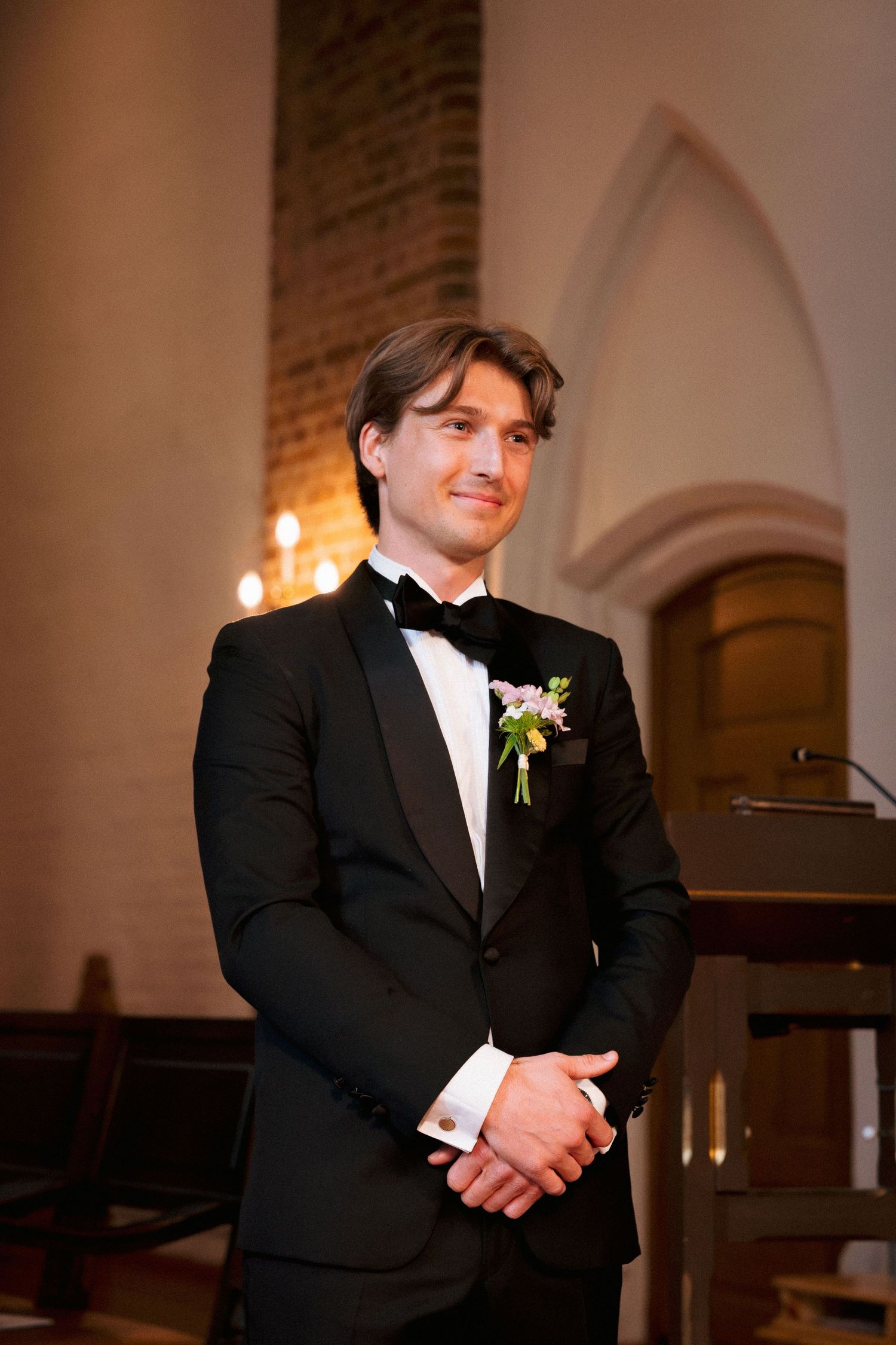 Groom in black tuxedo with floral boutonniere standing at church altar smiling.