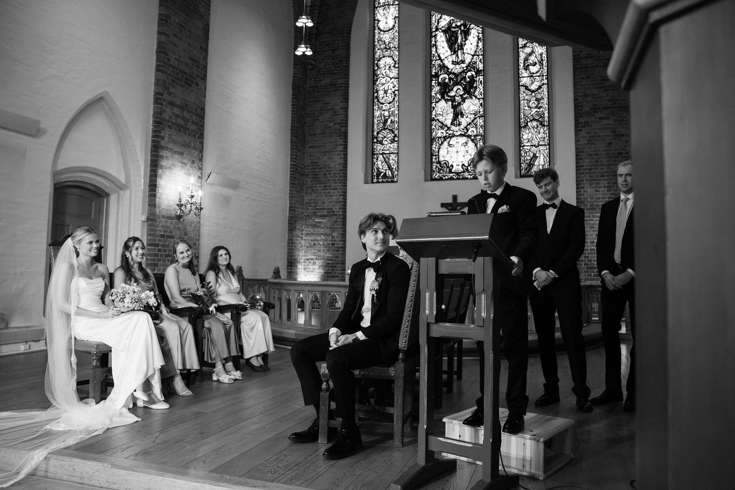 Black and white wedding ceremony photo inside a church with bride, groom, and guests near stained glass windows.