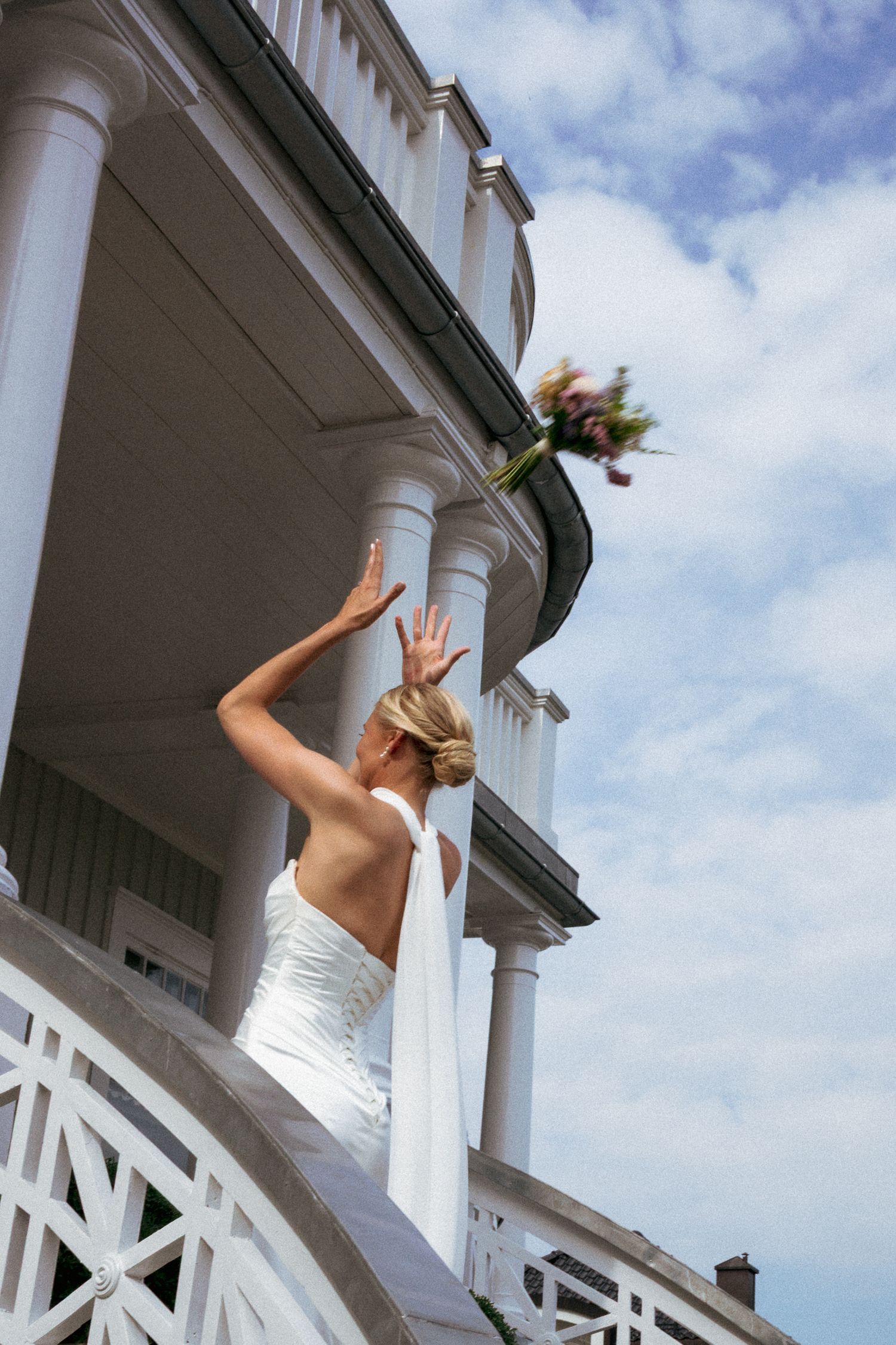 Bride in white backless gown tossing bouquet from elegant white mansion porch steps on wedding day.