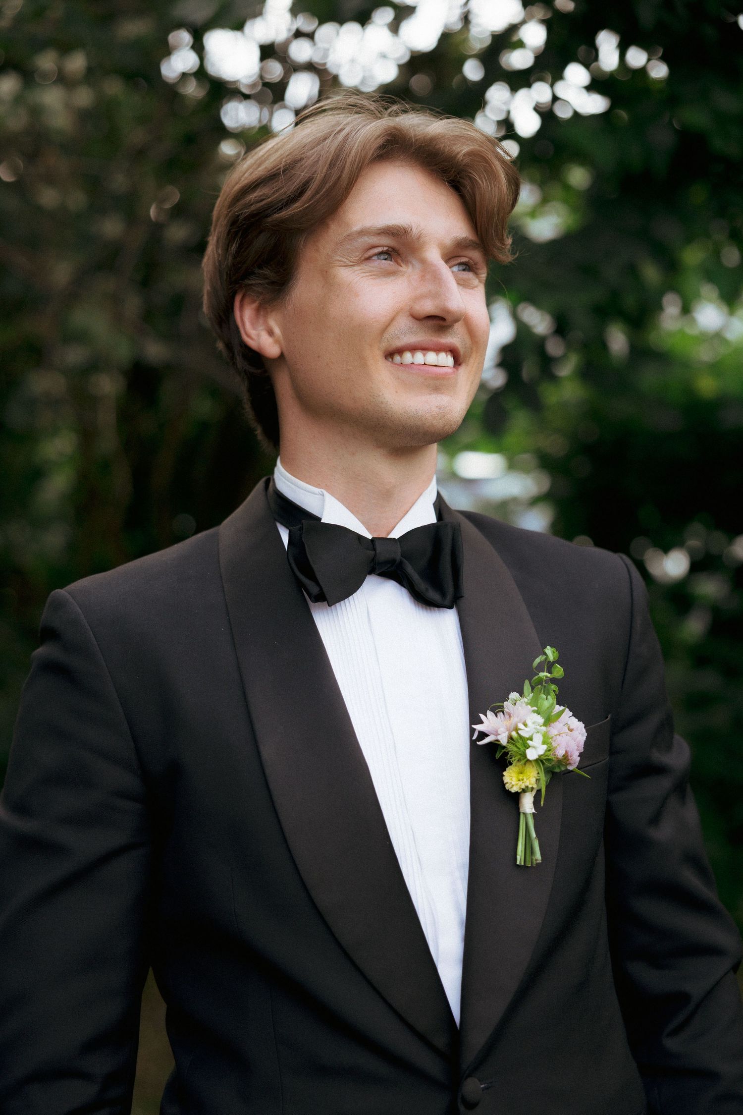Smiling groom in black tuxedo with floral boutonniere standing outdoors among green trees.