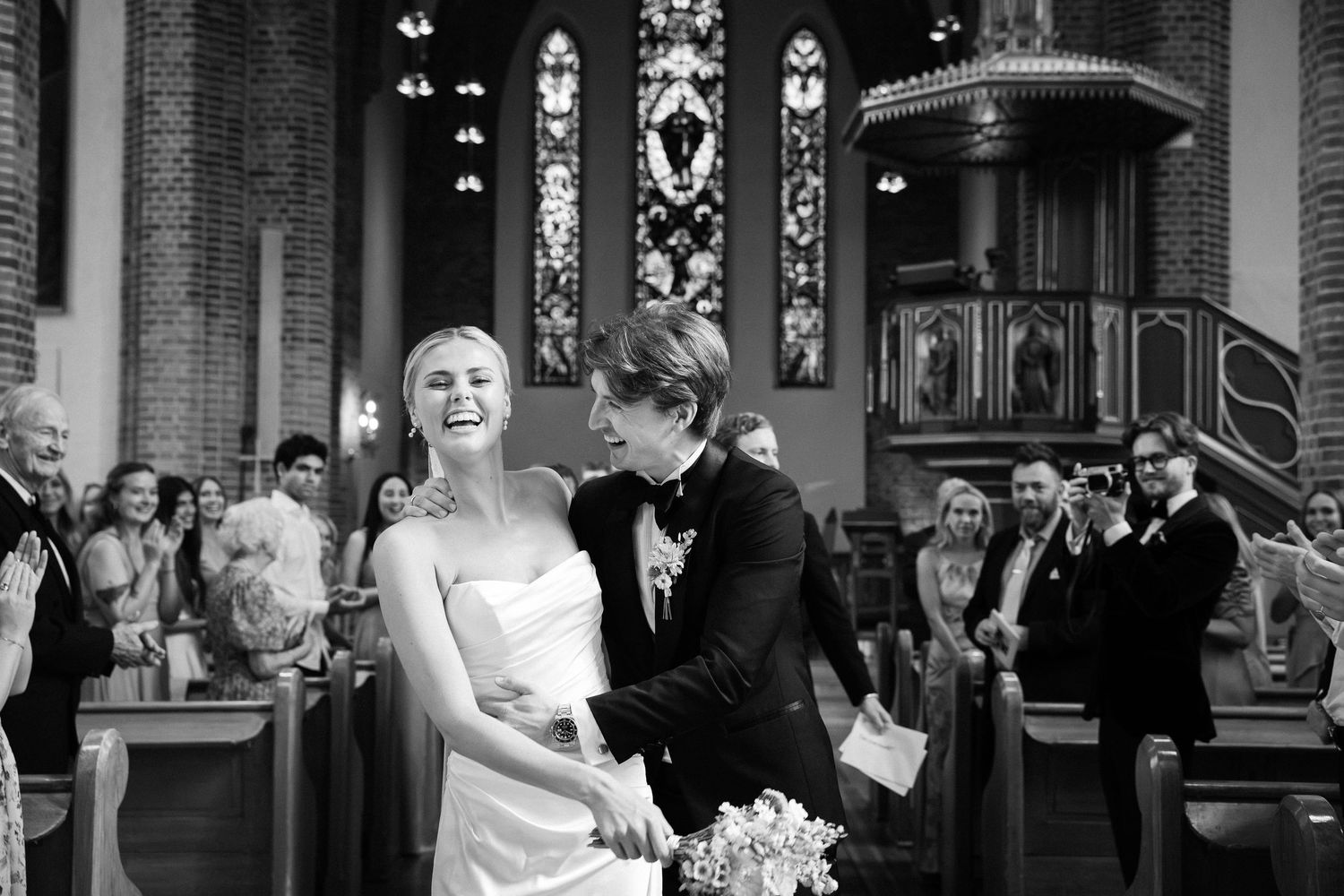 Happy bride laughs with groom during wedding ceremony inside elegant brick church with stained glass.