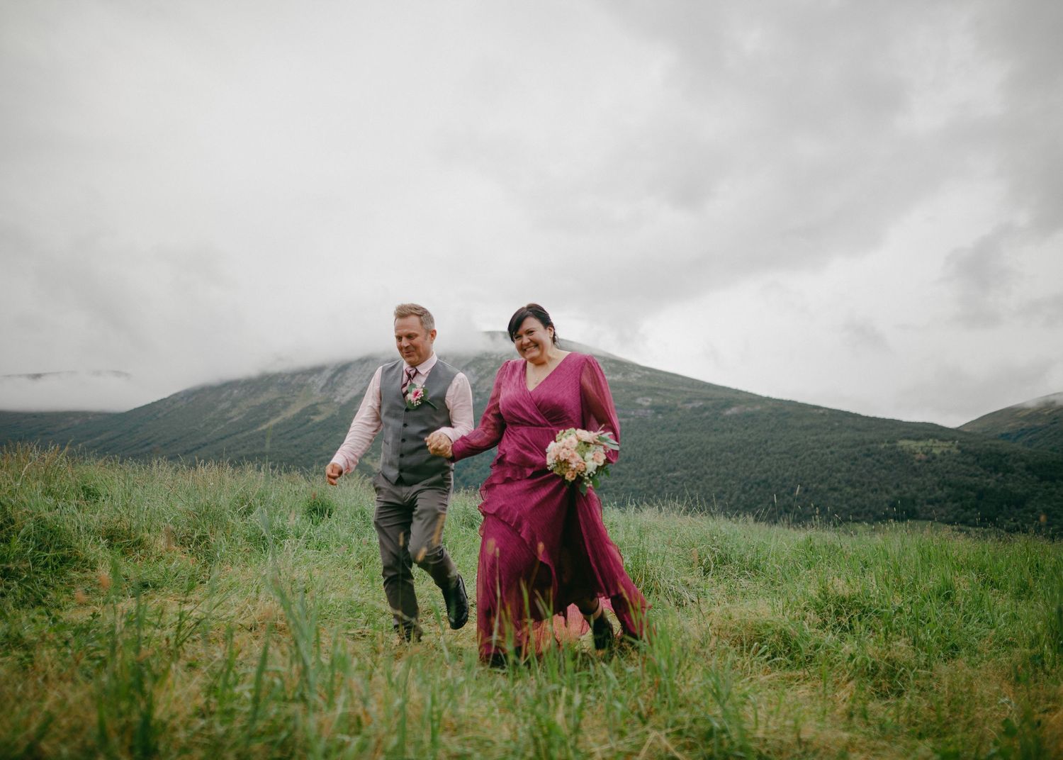 Bride in magenta dress and groom walking through green mountain meadow on wedding day.