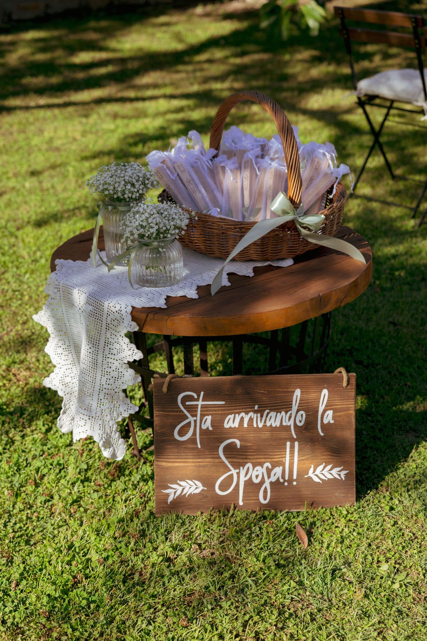 Rustic wooden wedding sign reading Sta arrivando la Sposa with wicker basket and white flowers on lace-covered table.