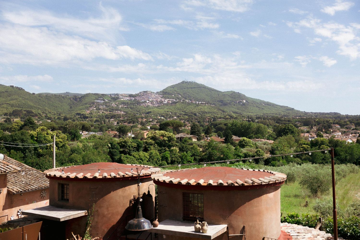 Panoramic view of a Mediterranean hillside village with terracotta rooftops, lush greenery, and a mountain backdrop under a blue sky.