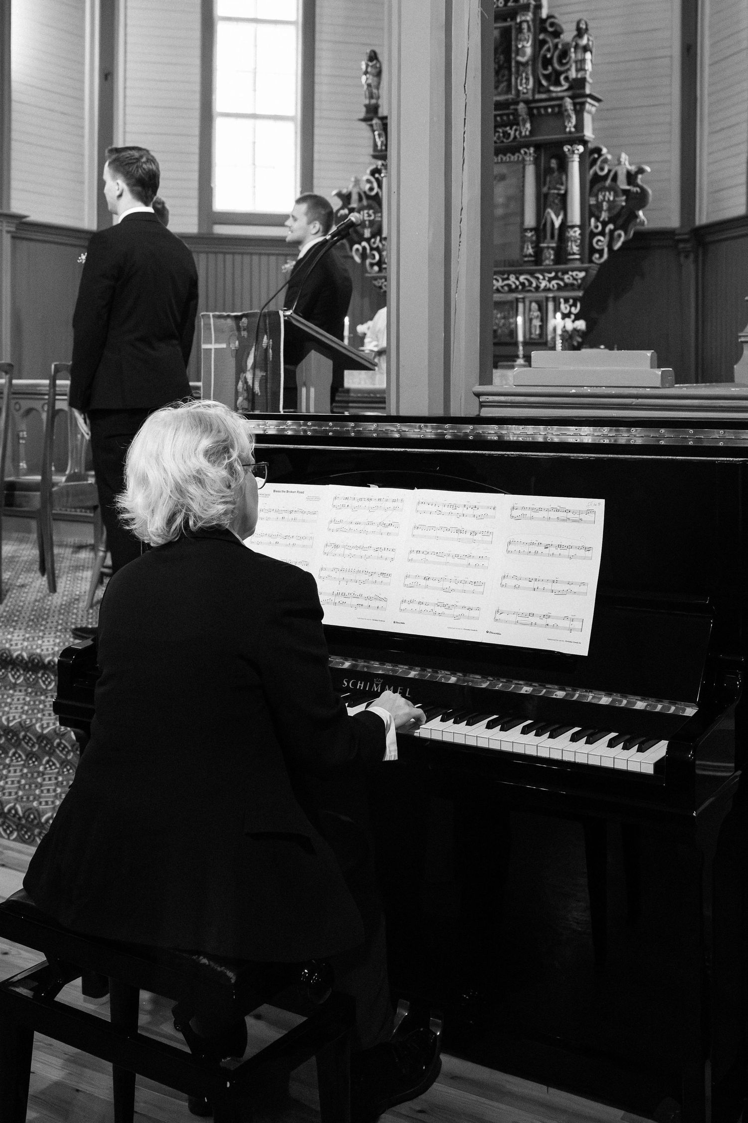 Black and white photo of elderly woman playing grand piano at church wedding ceremony with sheet music.