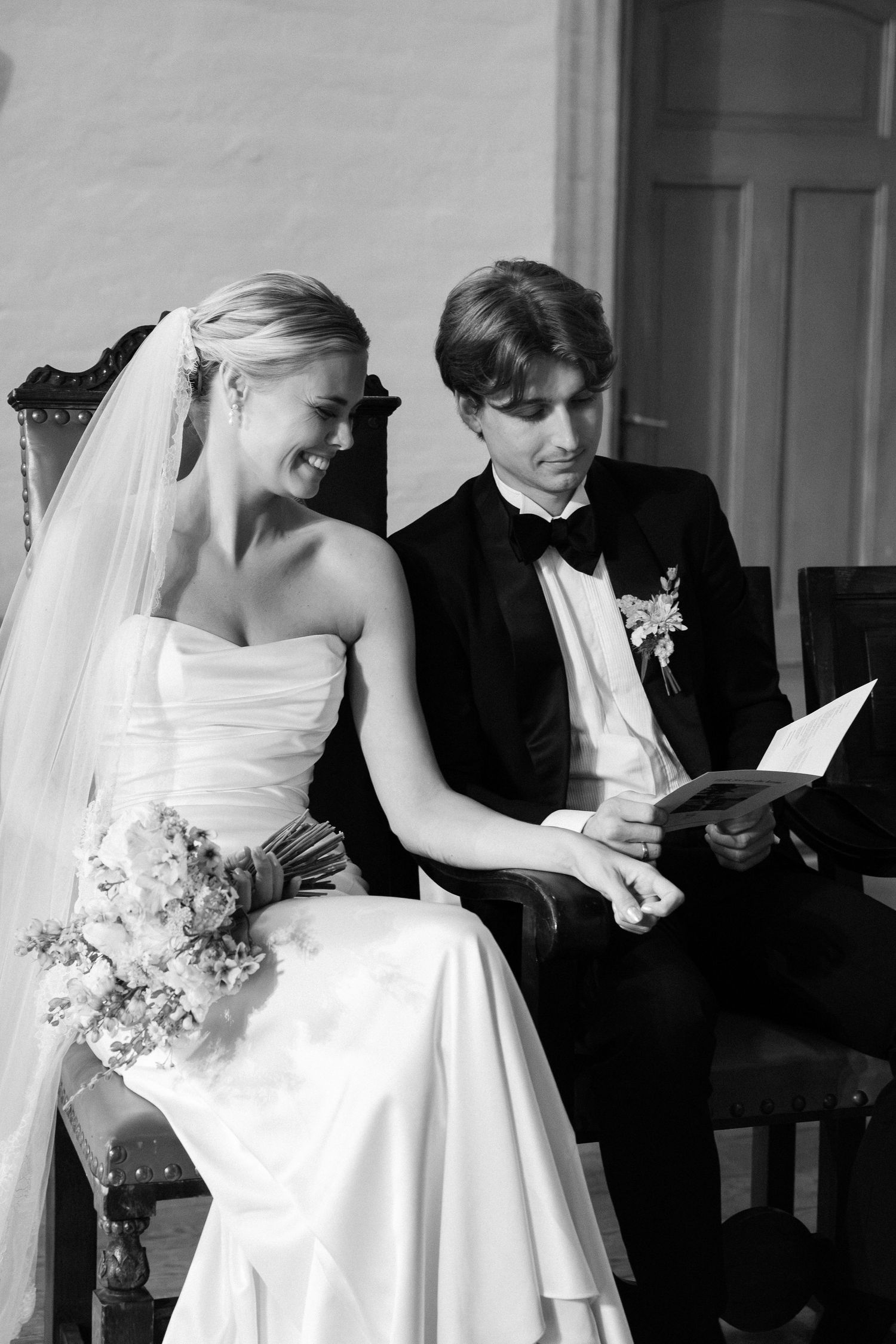 Bride and groom sitting together during wedding ceremony, bride smiling in strapless gown with veil and bouquet.