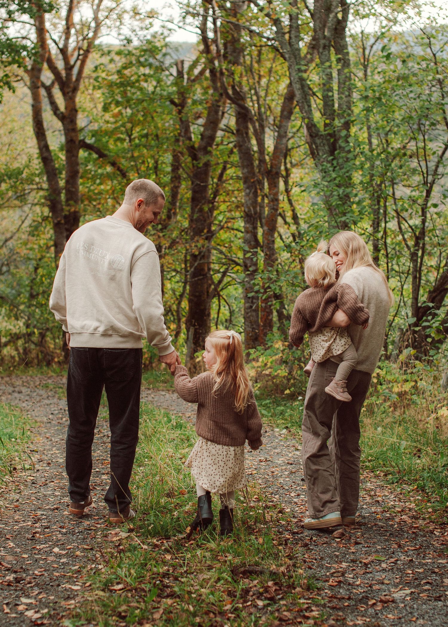 Family of four seen from behind on a forest path in autumn, parents holding children amid colorful fall foliage.