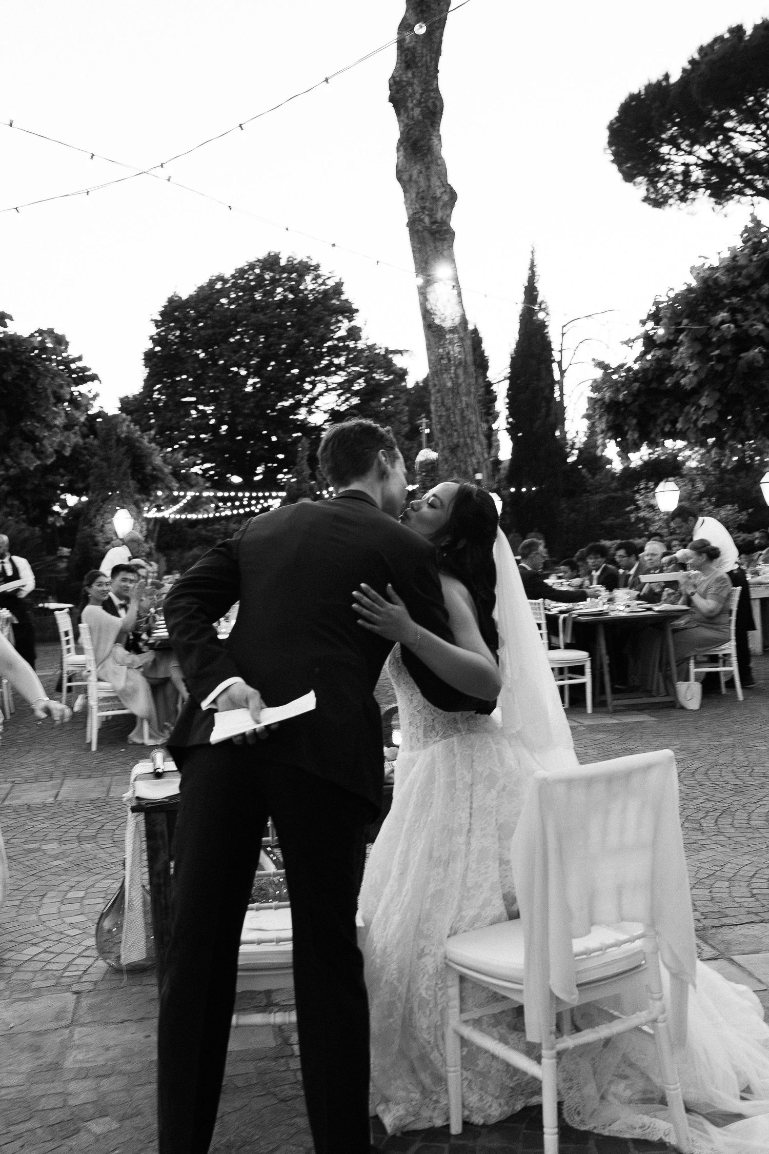 Black and white photo of bride and groom sharing first kiss at outdoor evening wedding reception.