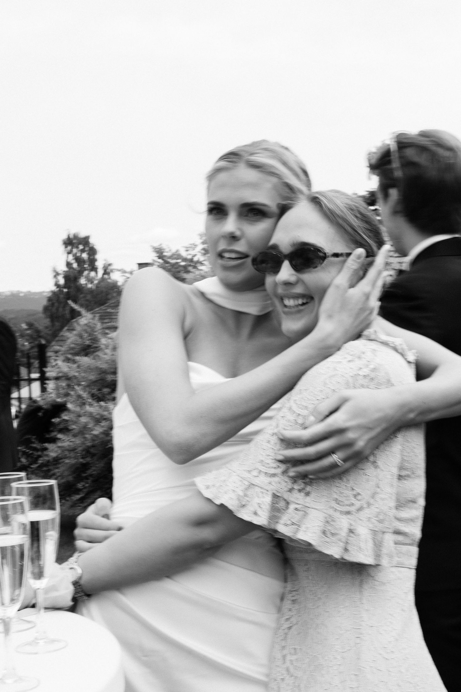Bride in white strapless gown hugging female guest wearing sunglasses at wedding reception.