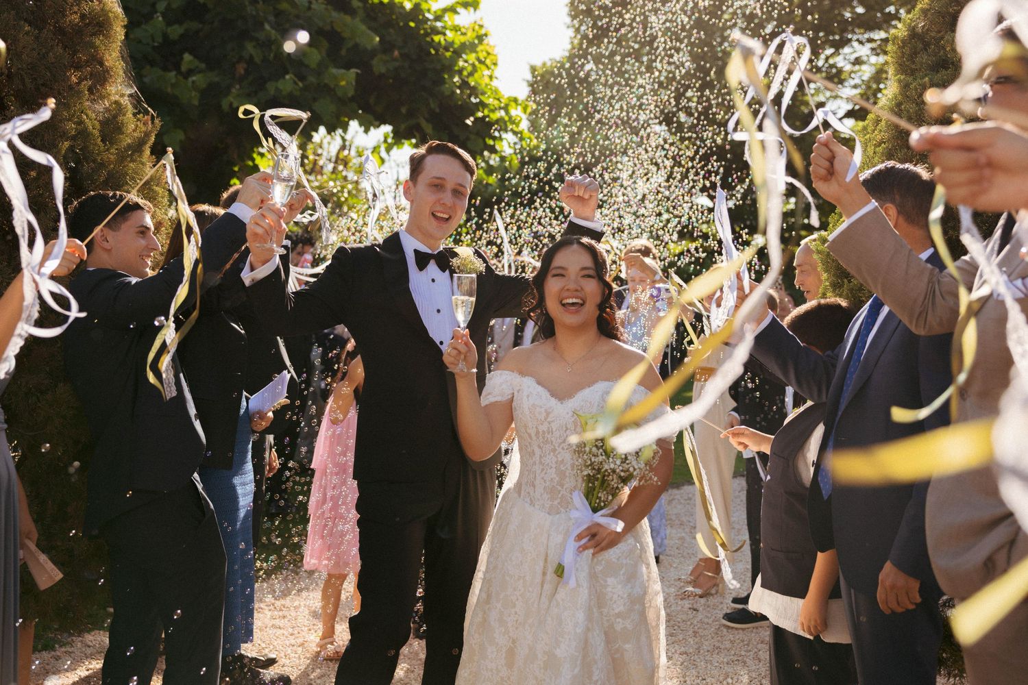 Joyful newlyweds walk through a confetti and ribbon sendoff surrounded by cheering guests at an outdoor wedding celebration.