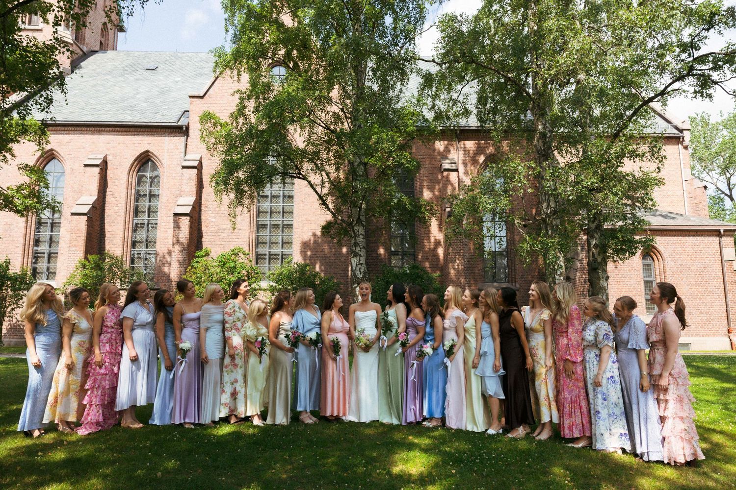 Large wedding party of bridesmaids in colorful pastel gowns pose together outside a historic brick church.