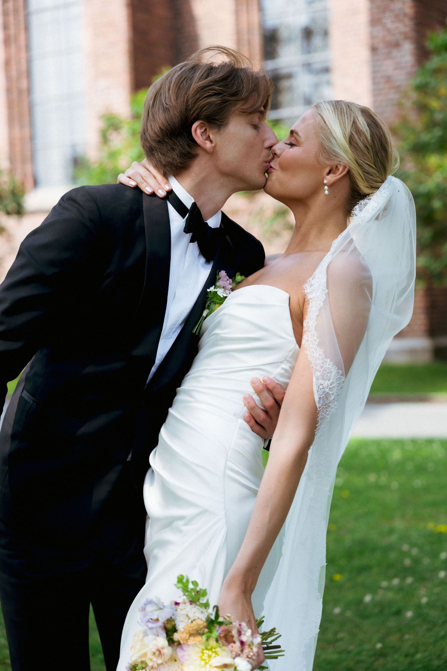 Bride and groom sharing a kiss outdoors, bride in white gown holding floral bouquet with veil.