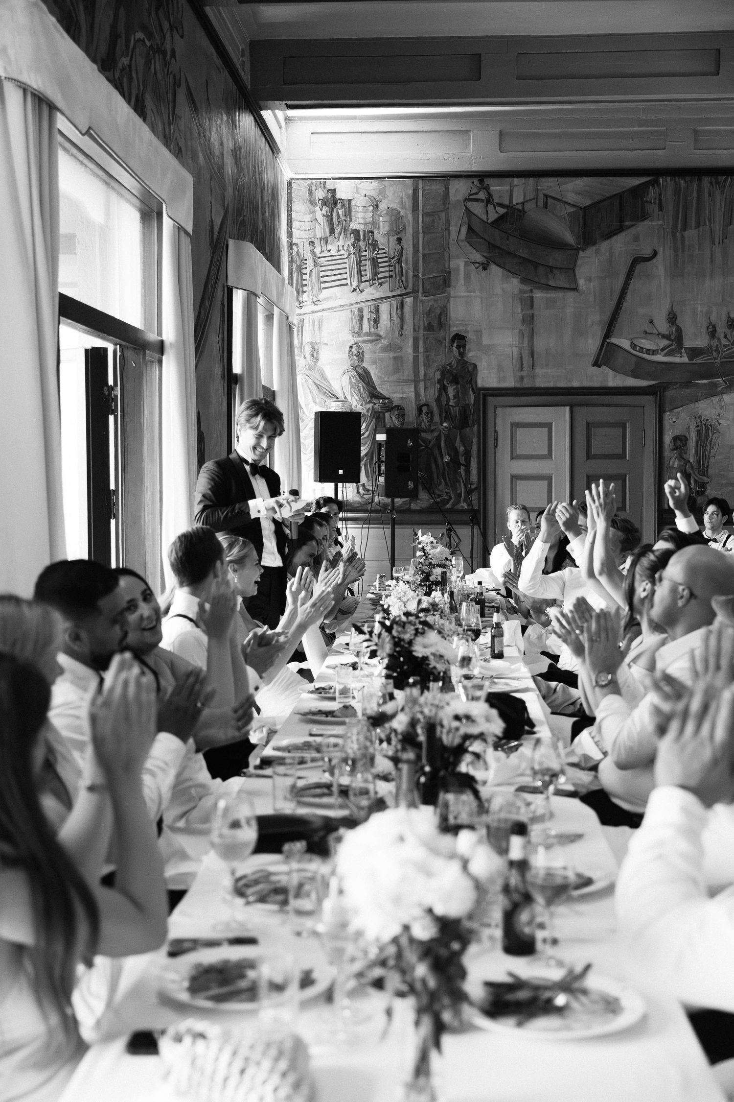 Wedding guests raise hands and applaud during lively reception dinner speech in elegant decorated hall.