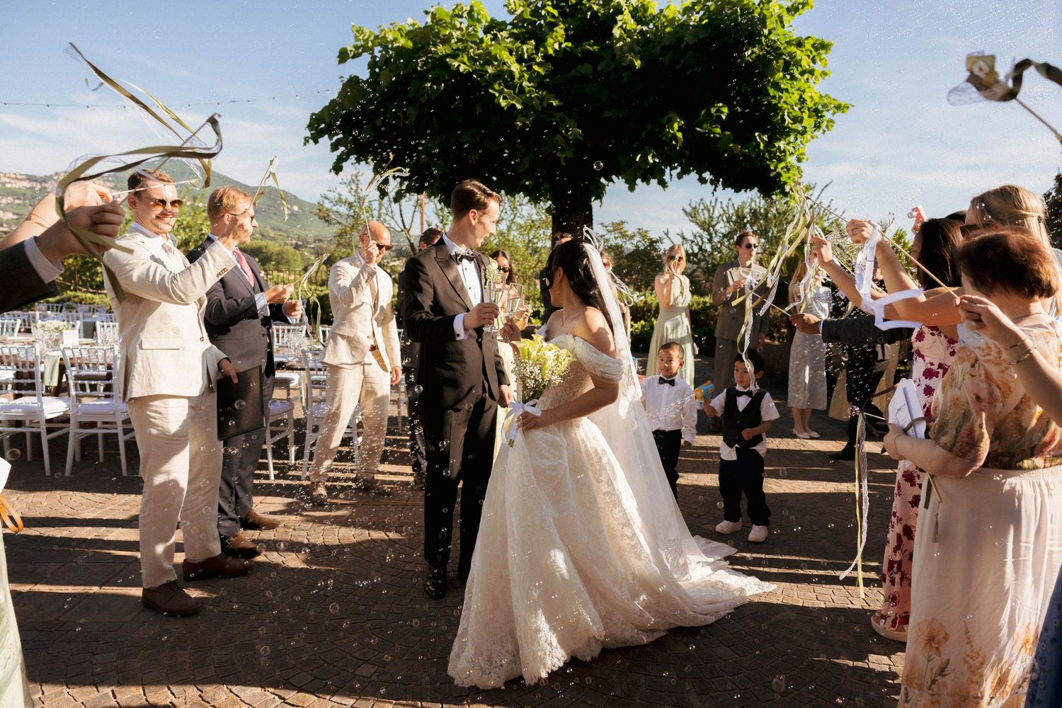 Bride and groom walk through cheering guests waving ribbons at outdoor wedding ceremony with mountain views.