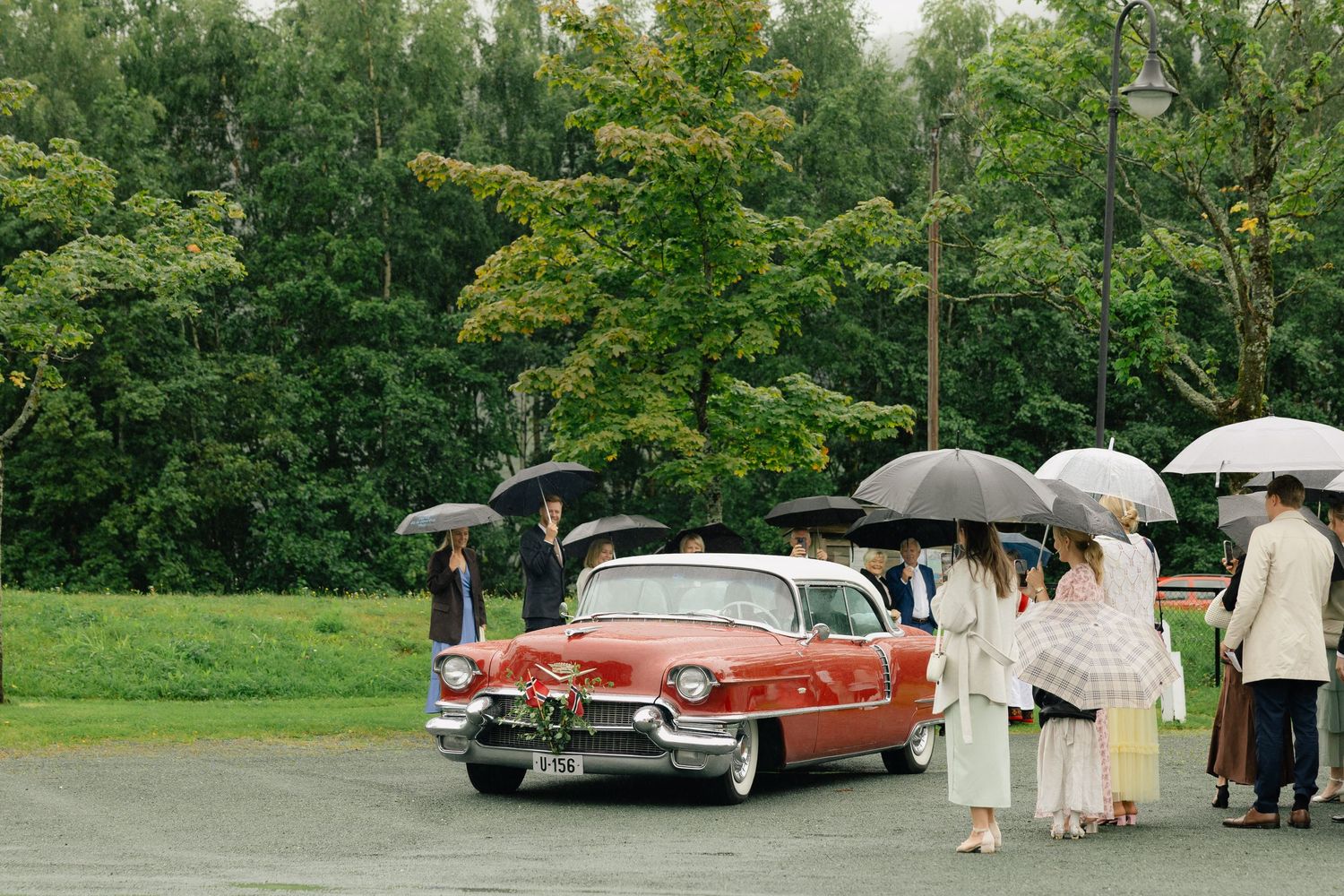 Wedding guests with umbrellas gather around a classic red vintage car on a rainy day outdoors.