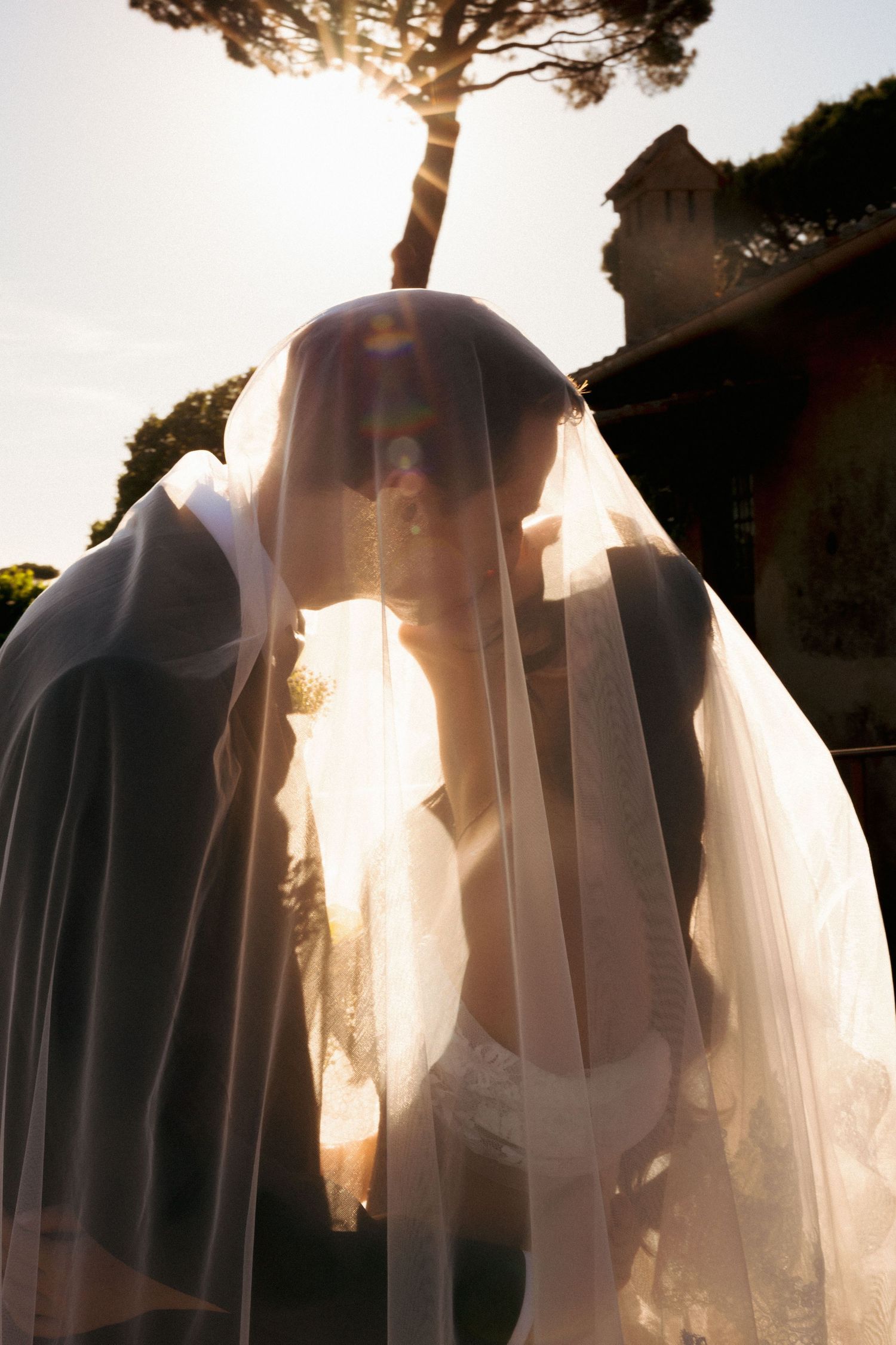 Bride and groom sharing a kiss under a flowing wedding veil with golden sunlight in an outdoor setting.