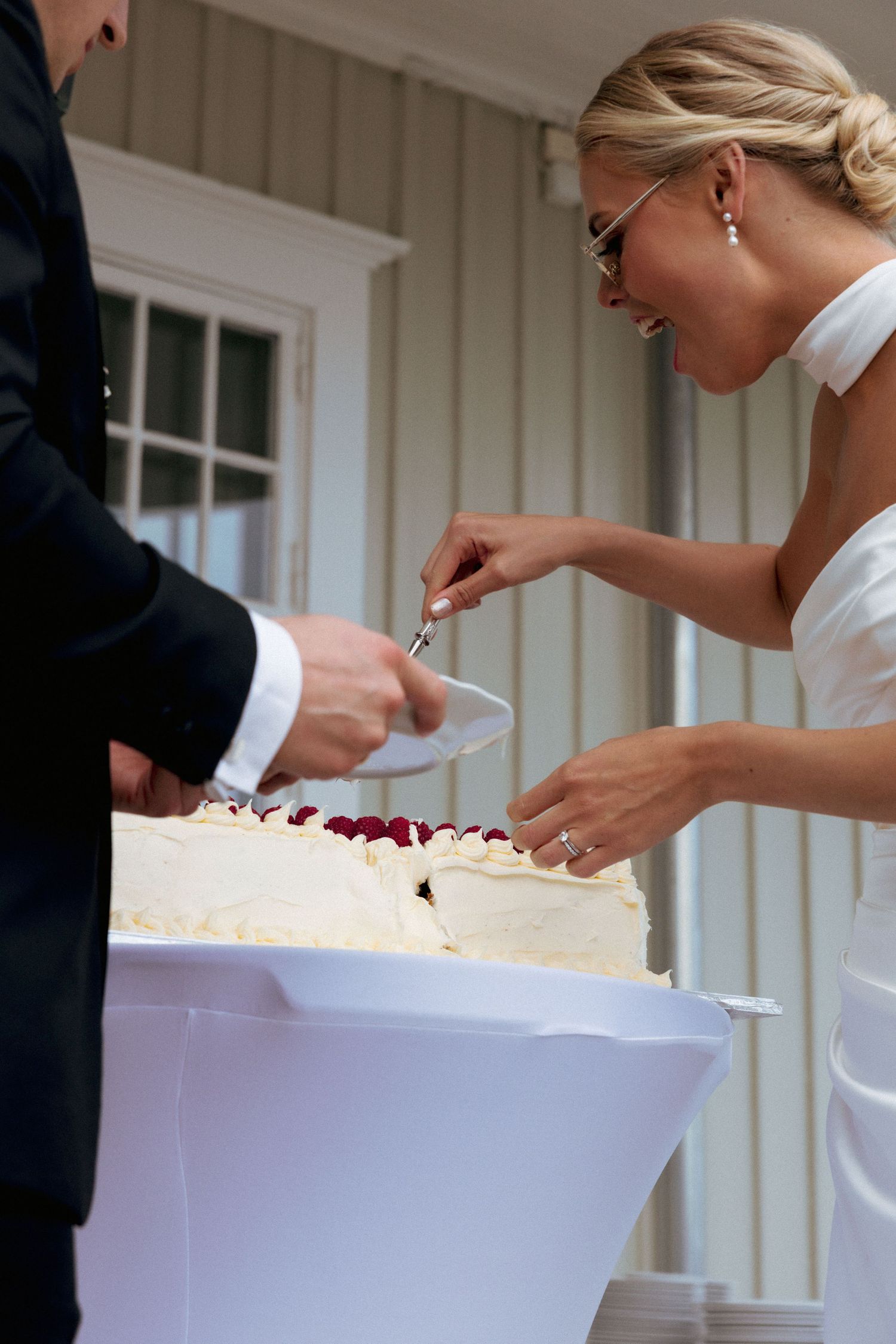 Bride in white strapless gown laughing while cutting wedding cake with groom at outdoor reception.