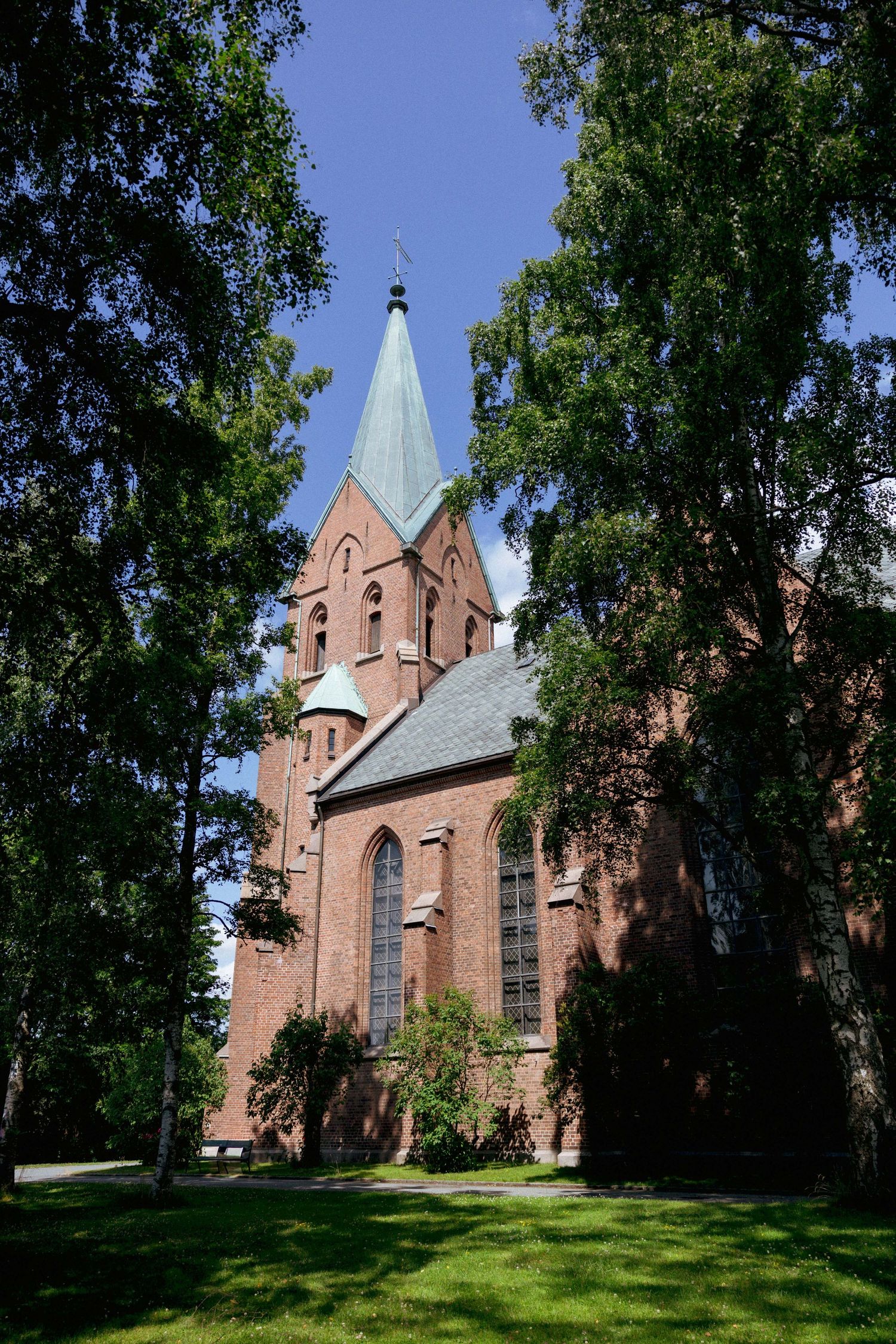 Red brick church with green copper spires surrounded by lush birch trees under a clear blue sky.