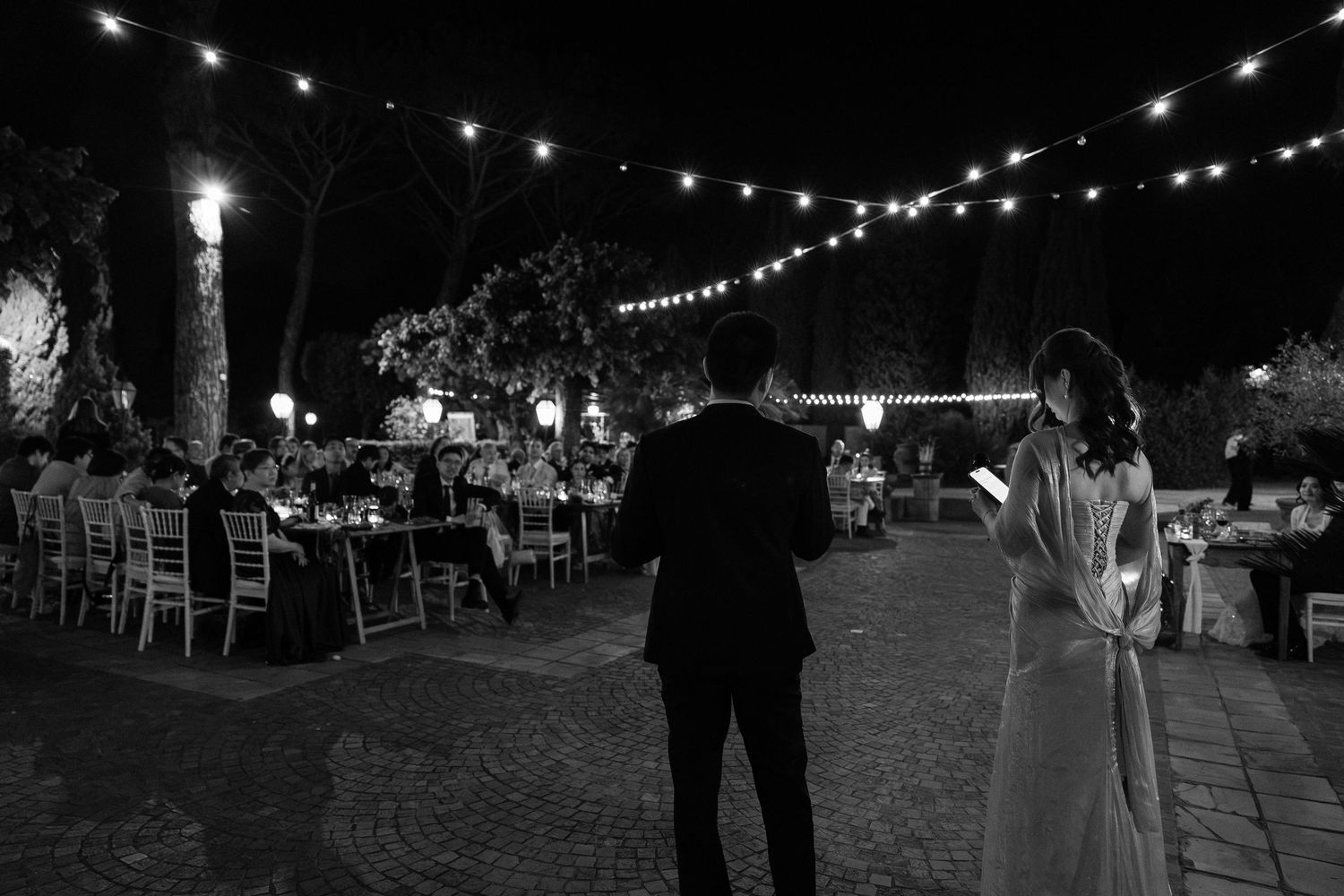 Black and white night wedding reception with string lights, guests dining outdoors as couple stands together.