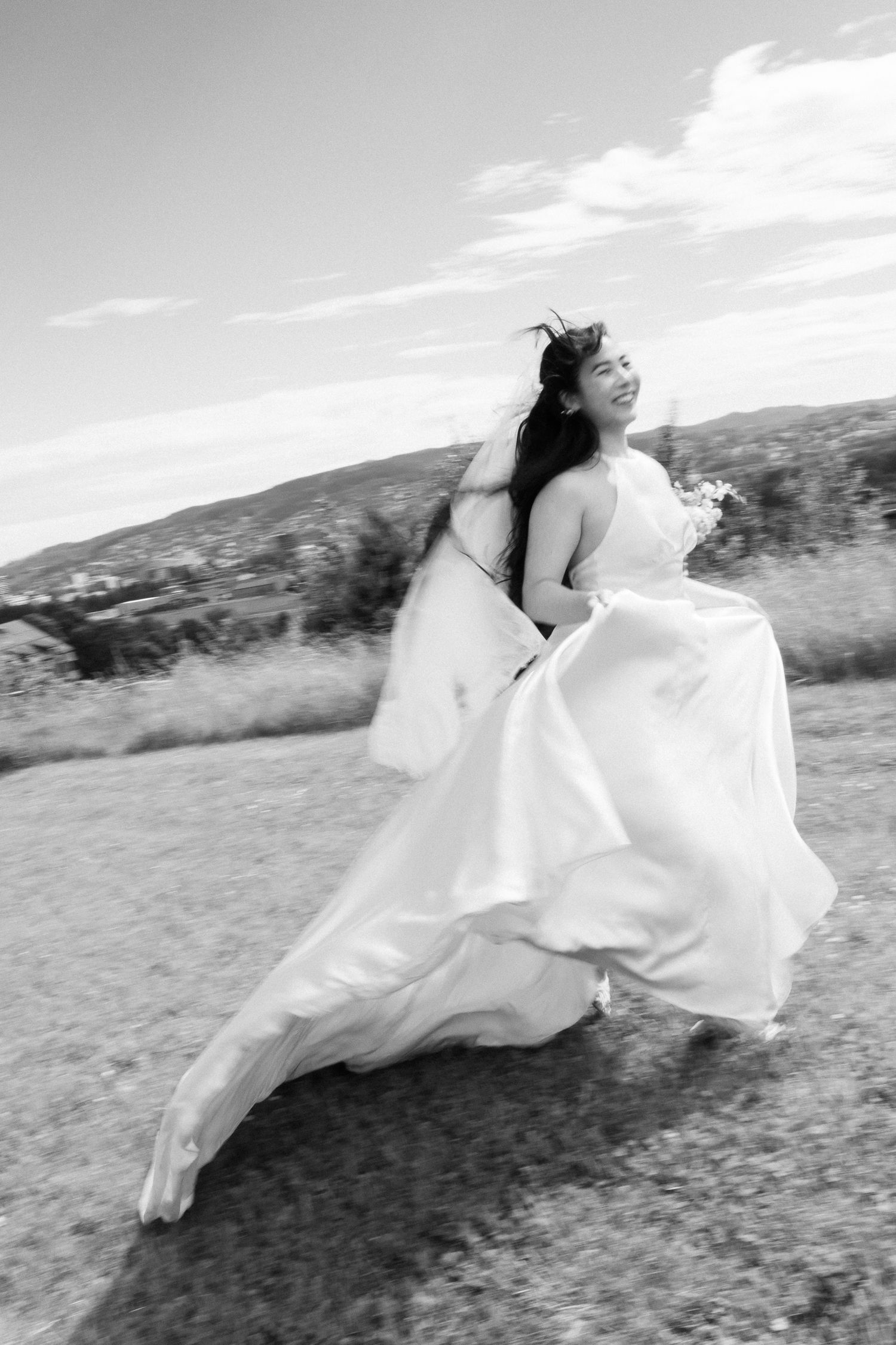 Black and white photo of a joyful bride in a flowing white gown running outdoors on her wedding day.