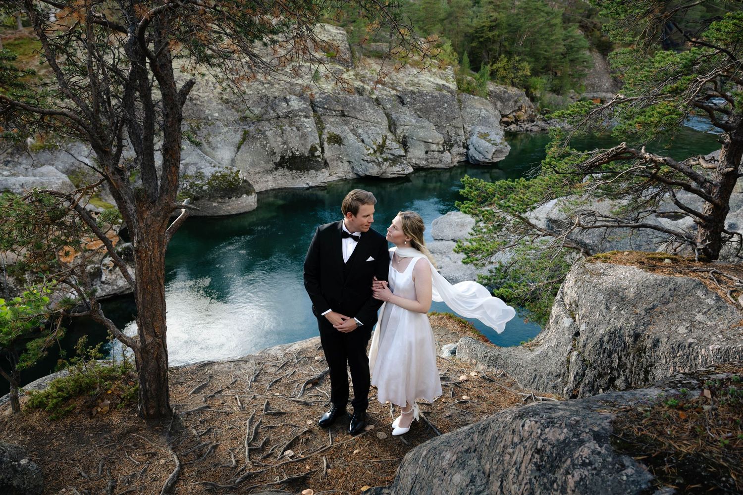 Bride and groom stand on rocky cliff overlooking turquoise river, sharing romantic moment in scenic outdoor wedding setting.